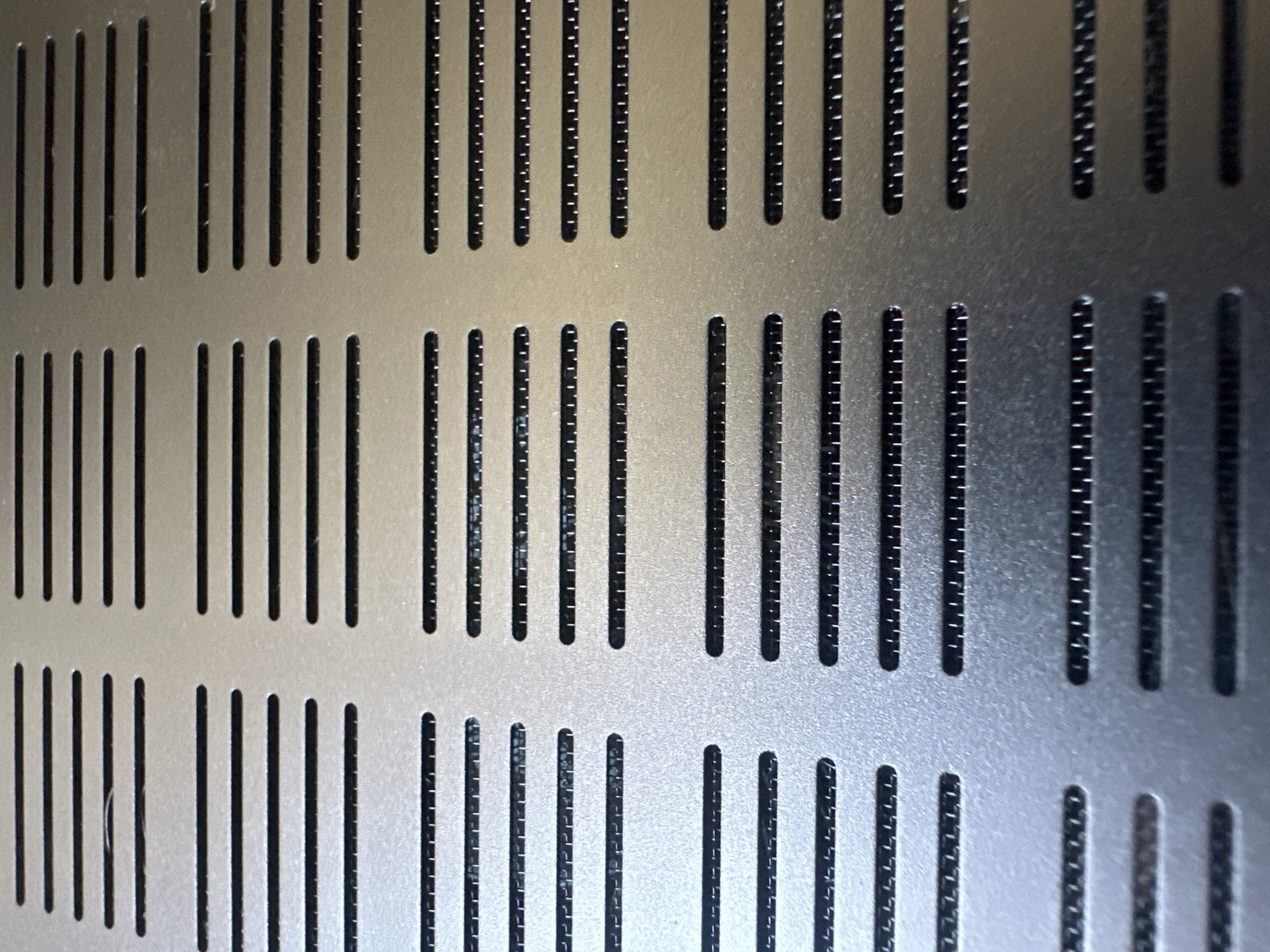 The image shows a close-up of a perforated metal surface, likely a ventilation grille. The surface is silver-grey with a brushed texture and features horizontal slots arranged in a grid pattern. Beneath some of the slots, a fine mesh can be seen, suggesting a filter or barrier. The lighting creates subtle shadows within the perforations, highlighting the depth and texture of the material. The image is a detail shot and does not provide any contextual information about its location in Uzhhorod, Ukraine, or any other environmental details.