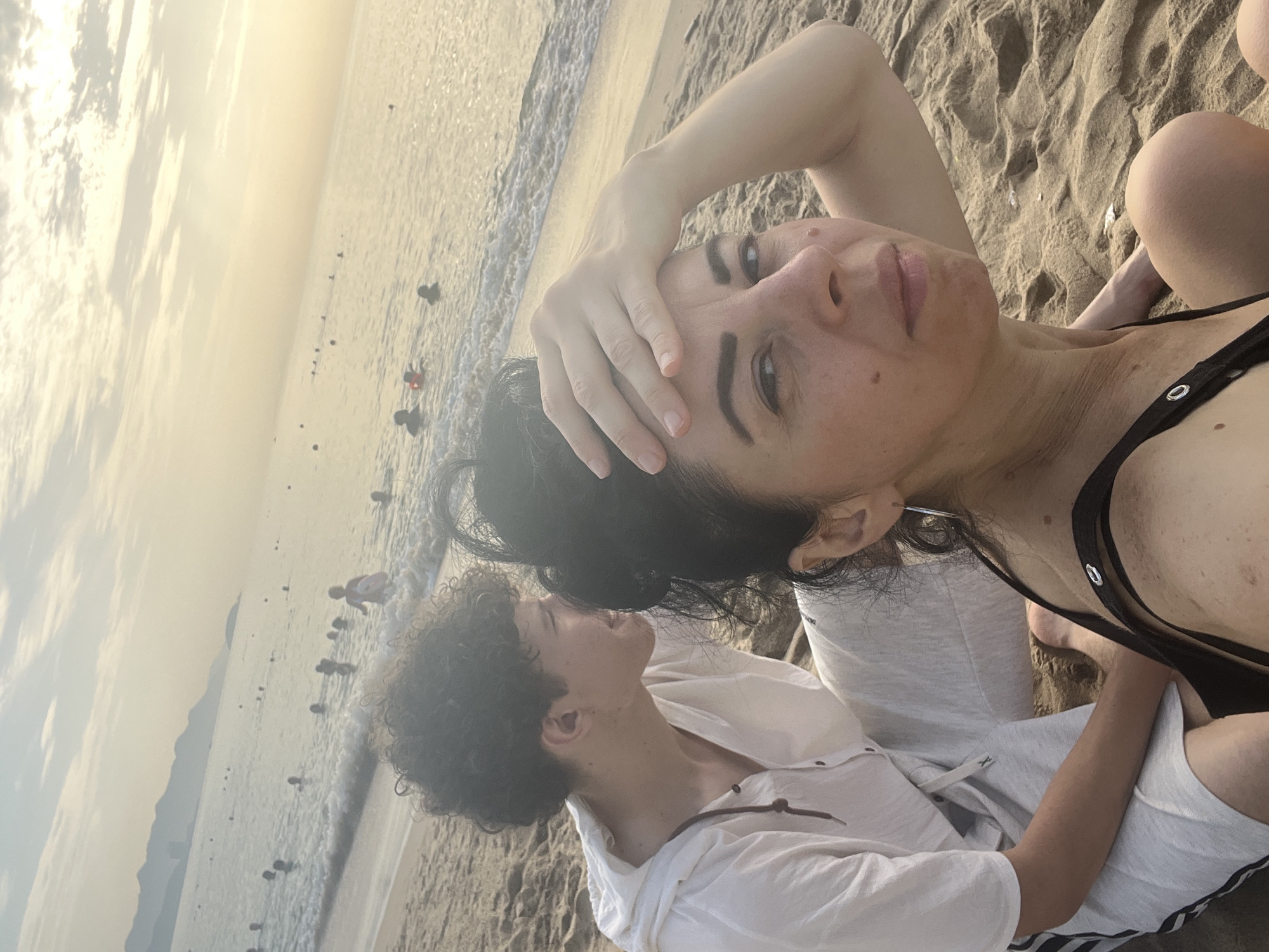 This image was taken on a beach in Nha Trang, Vietnam, at sunset. In the foreground, a woman with dark hair is holding her hand to her forehead, looking towards the camera. She is wearing a black bikini top. To her left, a young man with curly dark hair, wearing a white hoodie, is sitting beside her. The background shows the ocean with several people swimming and a hazy coastline with mountains. The sky is a pale orange and yellow, characteristic of a sunset. The sand is light brown.