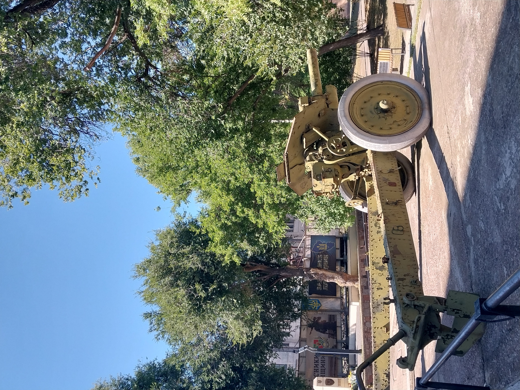 The image shows an anti-tank gun in an outdoor setting, likely a park or memorial site, in the city of Dnipro, Ukraine. The gun is painted in an olive-drab color and appears to be a static display, possibly part of a war memorial, given the presence of a monument in the background. It is positioned on a paved surface, with trees and blue sky forming the backdrop. The gun has a long barrel, a wheeled carriage with large white tires, and a distinctive recoil mechanism. The surrounding area is well-maintained, with some benches visible in the distance. The overall impression is one of a historical exhibit displayed in a public space.