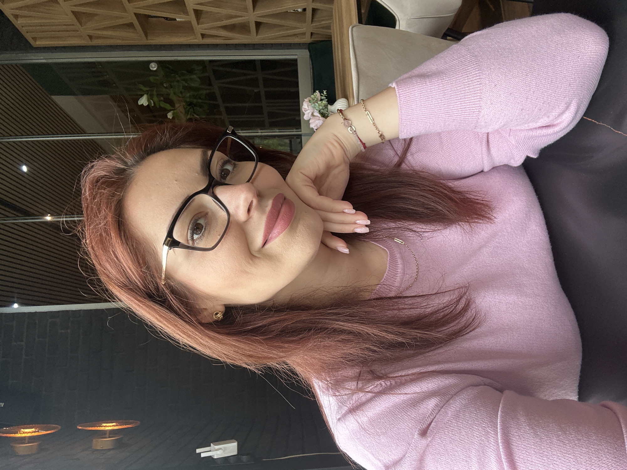 A person with reddish-brown hair wearing black-rimmed glasses and a pink sweater is posing for a selfie. She is resting her head on her hand. The location is unknown; the city of Montana, Bulgaria, cannot be determined from the image. The background shows a window, a shelf with a grid pattern, and furniture. The lighting suggests an indoor setting. There are two small table lamps on the bottom-left edge of the image.