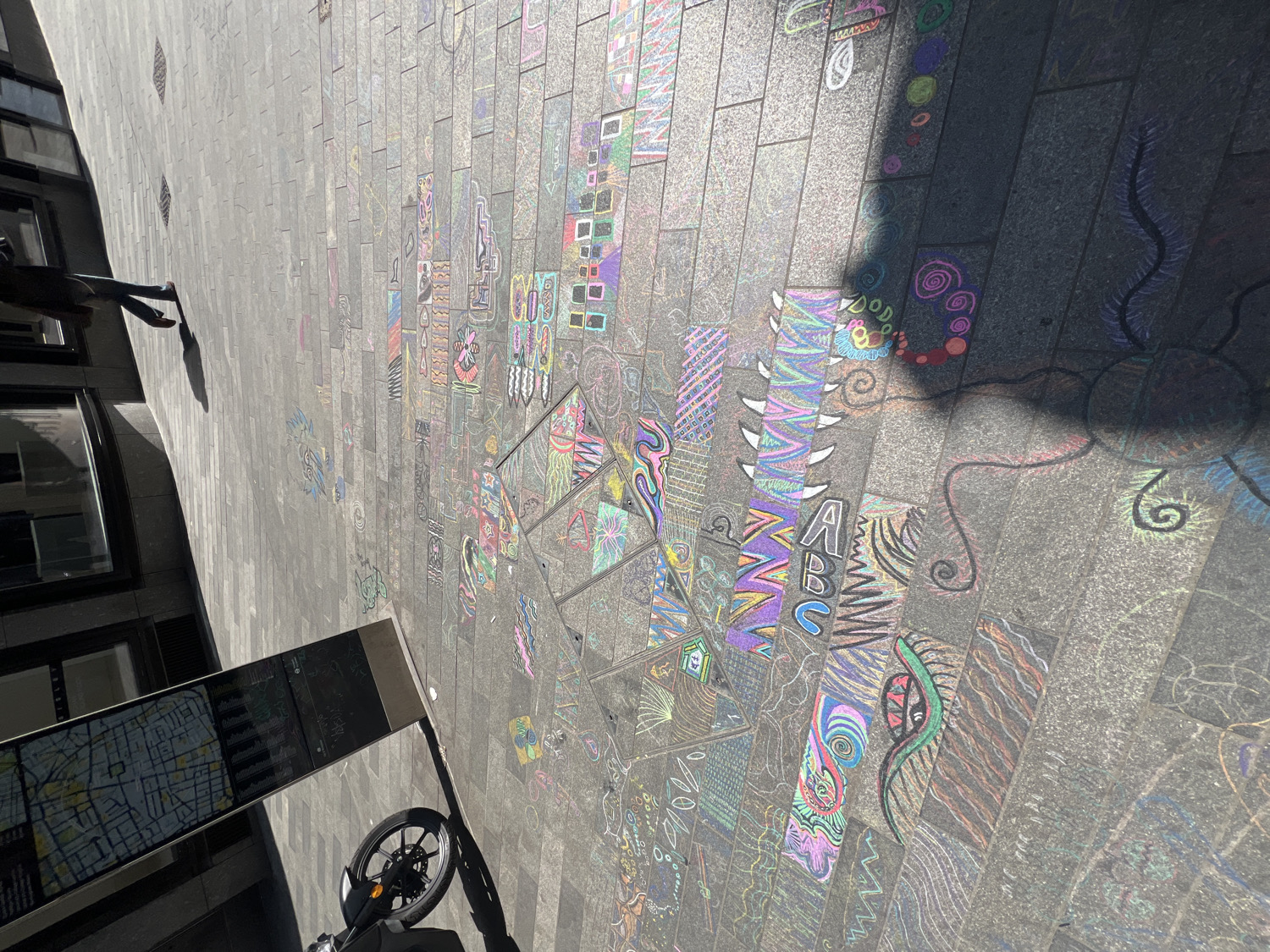 A vibrant, sun-drenched urban pavement, captured in a rotated, slightly elevated view, is extensively adorned with colorful chalk art. The dark grey rectangular paving stones serve as a canvas for a diverse array of drawings, featuring abstract patterns, intricate swirls, geometric shapes, and some designs resembling eyes or fantastical creatures. Visible text within the chalk art includes "ABC" and "DODO". The artwork utilizes a bright palette of purple, pink, blue, green, yellow, orange, and black, with designs often filling individual slabs or spanning across several.
On the left side of the image, modern buildings with reflective windows line the street. In the foreground, a street information board, displaying a map with city layouts, stands next to the front wheel and handlebars of a parked motorcycle or sturdy bicycle.
The scene is bathed in bright daylight, evidenced by distinct, long shadows cast across the pavement. These include the shadow of a person's upper body and legs in the upper left, and other shadows from the information board and vehicle falling onto the chalk art. This suggests either morning or late afternoon on a clear, sunny day, highlighting a lively public space where creative expression has taken place.