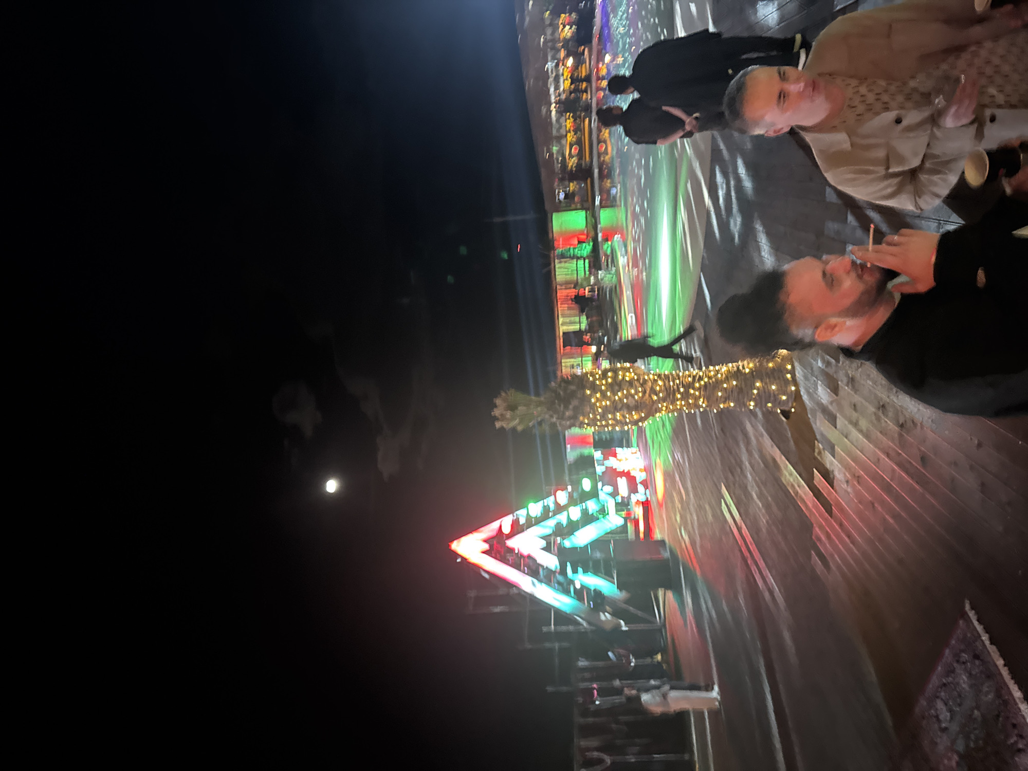 The image captures a nighttime scene in Parakar, Armenia, featuring several individuals on what appears to be an outdoor deck or patio. At the forefront, a man with dark hair is smoking a cigarette. Adjacent to him, another person is visible. Further back, more people are indistinctly grouped together. The setting is illuminated by a variety of lights. A Christmas tree made with red, green, and blue LED lights is visible in the background. Above this tree is a string of small yellow lights that extends overhead, and other buildings illuminated with lights are visible in the distance. To the left of the tree, a faint image of the moon is seen in the dark sky. The patio surface is made of what seems to be wooden planks, with a carpet partially visible at the edge.