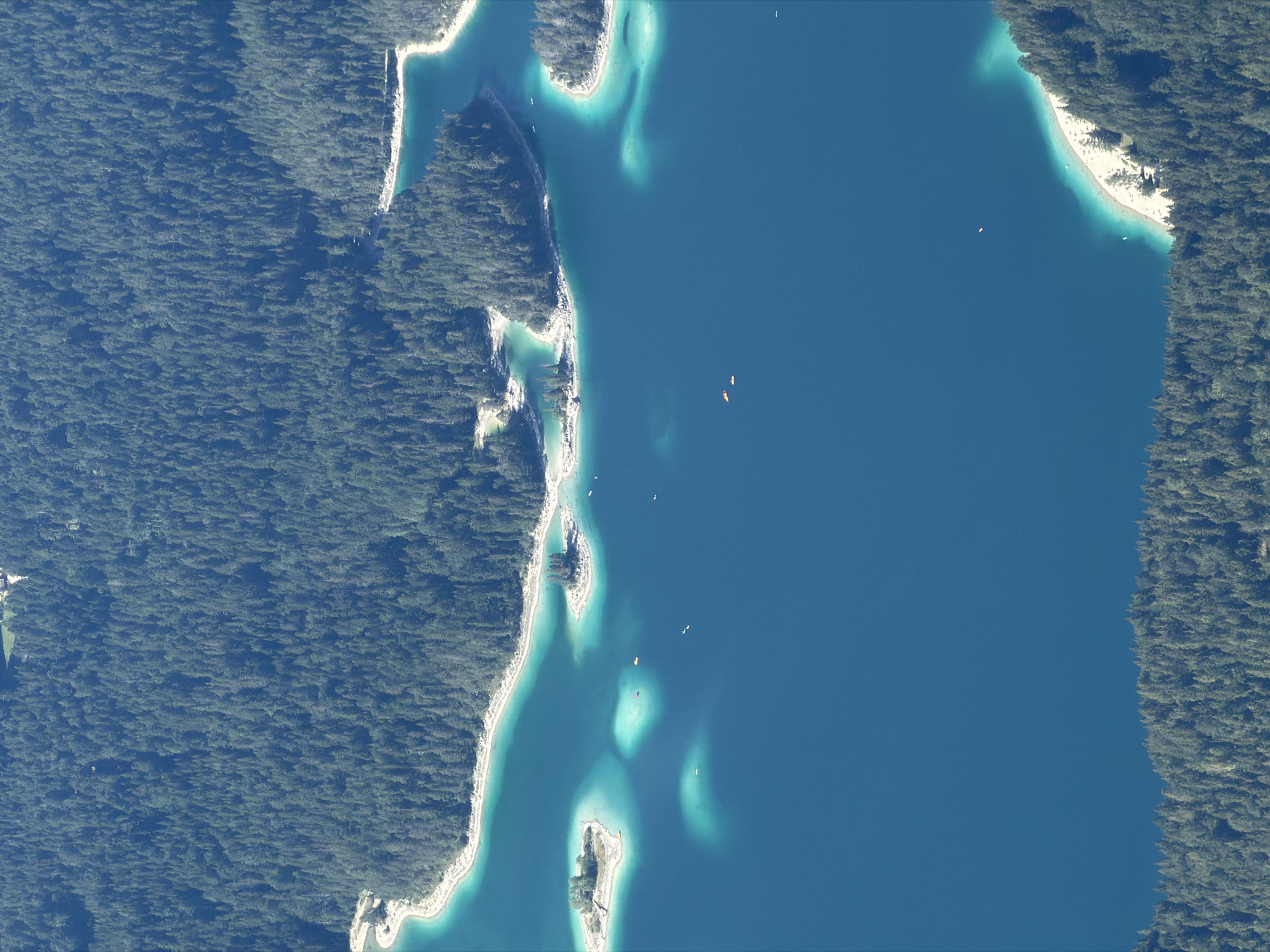 This aerial photograph captures a stunning view of the Eibsee, a picturesque lake located at the base of the Zugspitze mountain, near Garmisch-Partenkirchen, Germany. The lake's water exhibits a striking turquoise hue, particularly in the shallower areas and along the shoreline, contrasted by a deep sapphire blue in its depths. Several small, tree-covered islands dot the surface of the lake, adding to its scenic beauty.
Along the edges of the lake, a dense, dark green forest covers the surrounding hills and mountains. The forest is predominantly composed of coniferous trees, creating a textured canopy. The shoreline features a mix of rocky areas and sandy beaches, where a few people can be seen relaxing. Several small boats, including kayaks and possibly paddleboats, are scattered across the water, indicating recreational activity.
The overall scene is one of natural tranquility and scenic splendor. The image provides a clear, high-angle perspective, showcasing the interplay of water, land, and vegetation in this mountainous region of Bavaria.