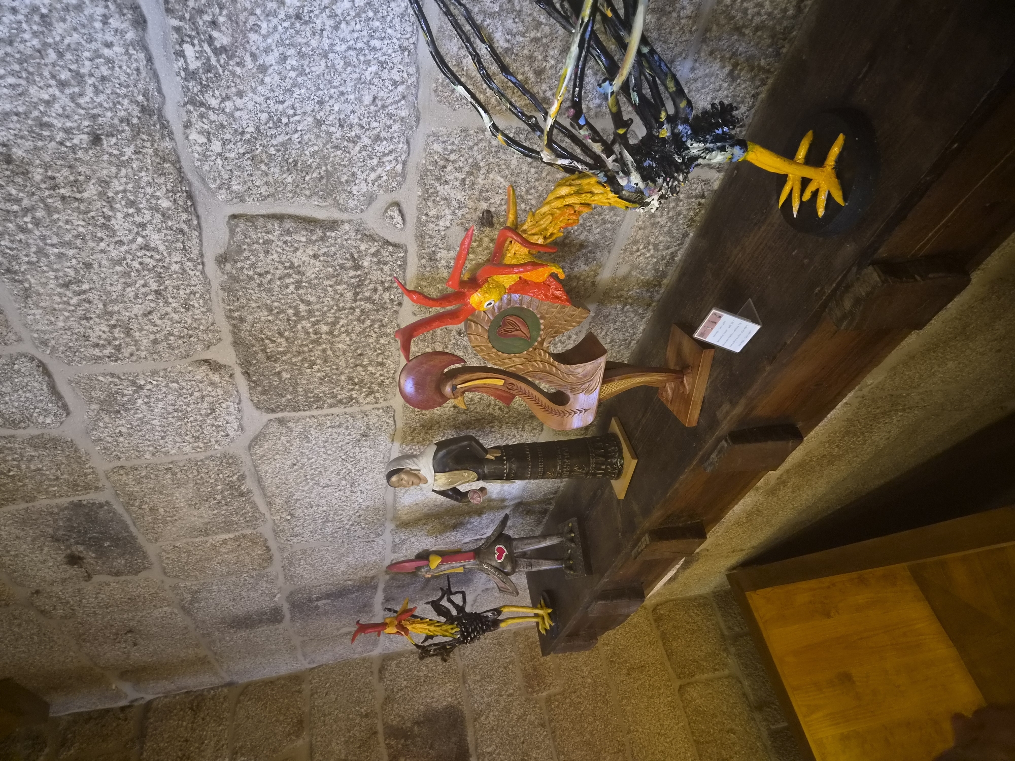 This image depicts a display of decorative ceramic figurines of roosters and a woman, situated on a wooden shelf against a rough stone wall. The figurines are colorful and varied in design, with several depicting stylized roosters and one a woman in traditional attire. The setting appears to be indoors, possibly a museum or a shop, given the display shelf and the stone architecture. The overall impression is one of folk art or cultural artifacts. This scene is located in Barcelos, Portugal, a city famous for its Roosters of Barcelos, a national symbol of Portugal, which are traditionally ceramic figurines.