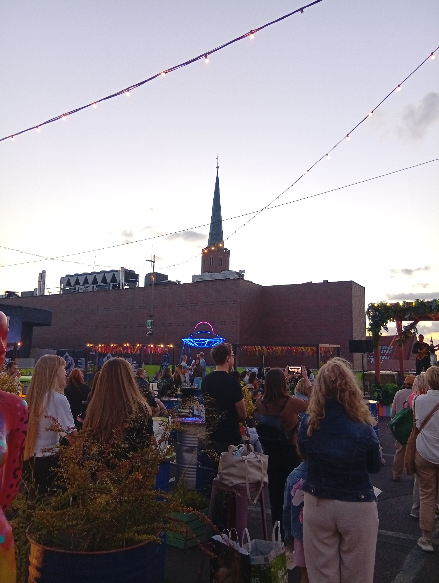 This image depicts an outdoor event at dusk, with a crowd gathered in front of a large brick building topped with a steeple. String lights are strung across the sky, and a neon UFO-like sculpture is visible in the background. A musician is playing a guitar on a raised platform adorned with greenery. People are standing and sitting, some holding phones to record. The scene suggests a festival or concert taking place in an urban setting. The presence of the steeple and brick building might indicate a historical area. It is possible this is the area of Wrocław, Poland.