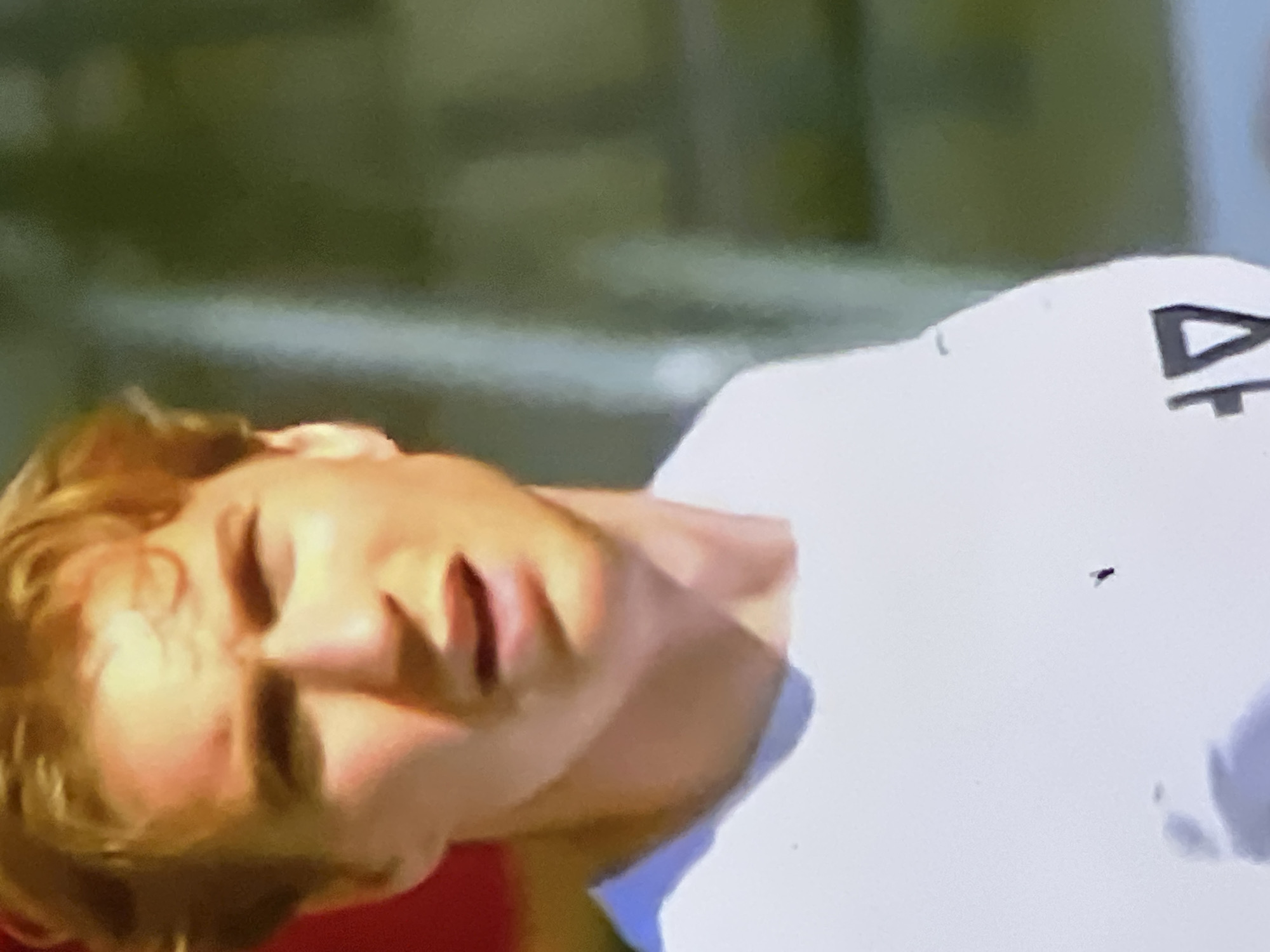 The scene, located in the city of Casablanca, Morocco, features a close-up of a young adult male. He has reddish-blonde hair and light skin, illuminated by strong, direct lighting from the viewer's left, creating defined shadows on his right cheek and under his chin. His mouth is slightly open, and his eyes are directed to the right side of the frame. He wears a plain white crew-neck t-shirt. A small dark speck is visible on the left side of his chest. On the lower right portion of the shirt, a partial dark logo or text is discernible, including a prominent triangular shape. The background is out of focus, predominantly dark green and grey, with a blurred red element visible to the far left.