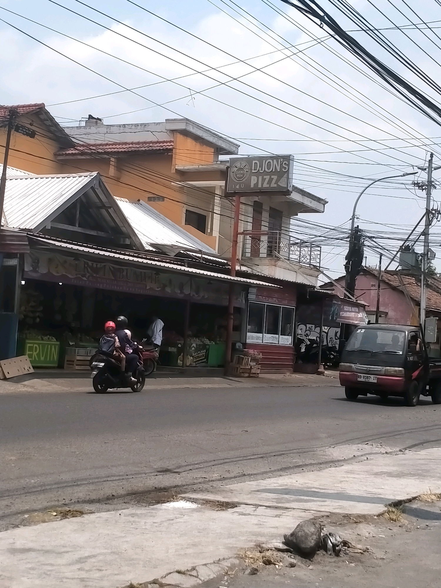 In the city of Kasihan, Indonesia, an overcast sky looms above a street scene. On the left, a building with an orange facade features a prominent sign for "DJON'S PIZZ." Below, a market stall displays various fruits and vegetables. Two people on a motorcycle are captured in motion on the road. To the right, a red and black pickup truck drives by, its license plate reading "AB 8187 IT." The scene is marked by a dense network of overhead electrical wires, typical of urban environments in Indonesia.