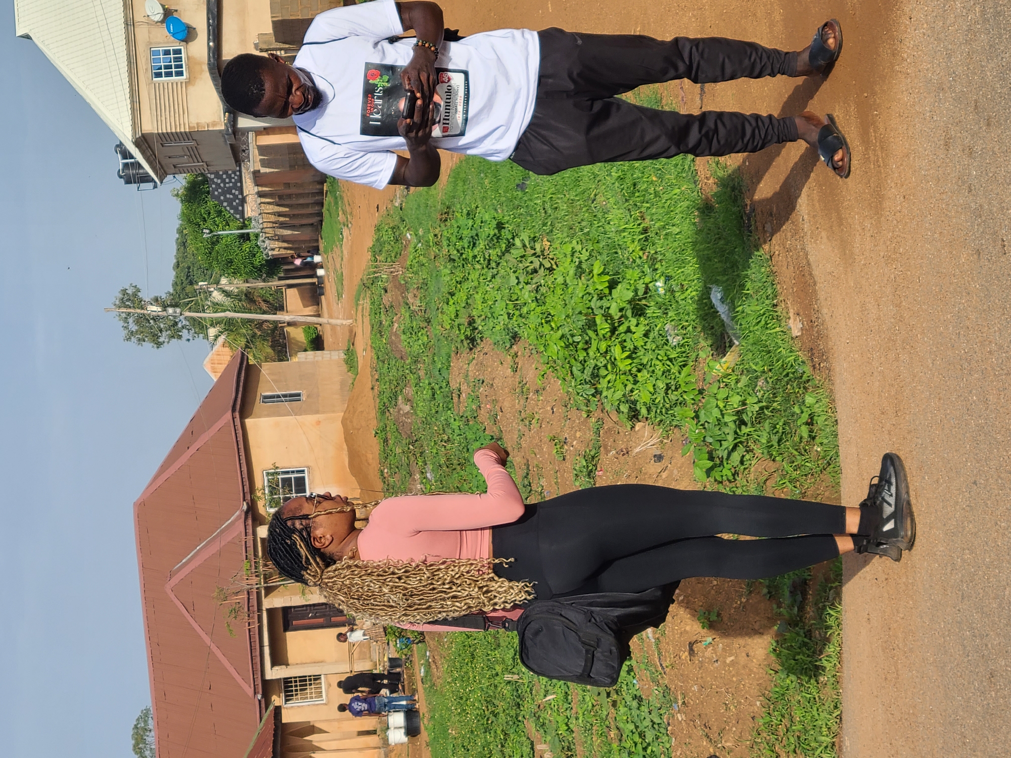 A full outdoor shot shows two people in the city of Unknown, Unknown. A woman with long blonde hair braided into twists, wearing sunglasses, a long-sleeved pink shirt, black leggings, and black sneakers stands on a dirt path. She has a black bag slung over her shoulder. Adjacent to her on the right is a patch of weeds. In the background is a tan building with a brown roof and barred windows.
A man stands to the upper left of the woman on a similar dirt path. He wears a white short-sleeved t-shirt, black pants, and dark sandals. He is holding a black phone in his hands, appearing to be texting or taking a photo.
Behind the man is a similar patch of weeds, and in the background, a beige building with a white roof and a blue satellite dish. The sky is clear and blue.