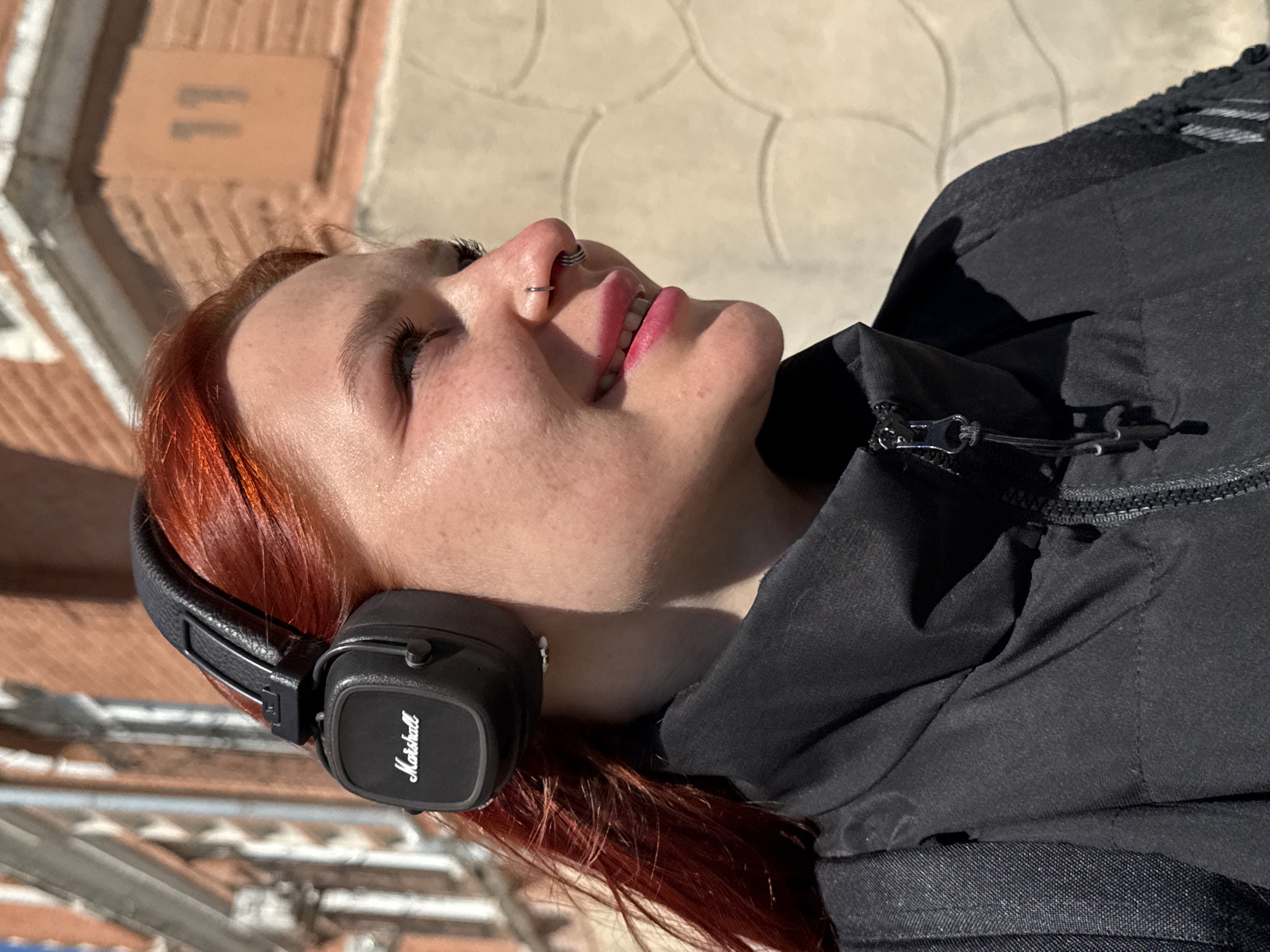 A young woman with red hair is depicted in a low-angle shot while wearing black Marshall headphones. She has a nose piercing, and her face is angled upward, revealing a pink lipstick shade. She is wearing a black zip-up jacket. The background appears to be an urban setting, likely a street in Rostov-on-Don, Russia, based on the architecture and paving stones, although without further context or metadata, identifying the exact location remains speculative.