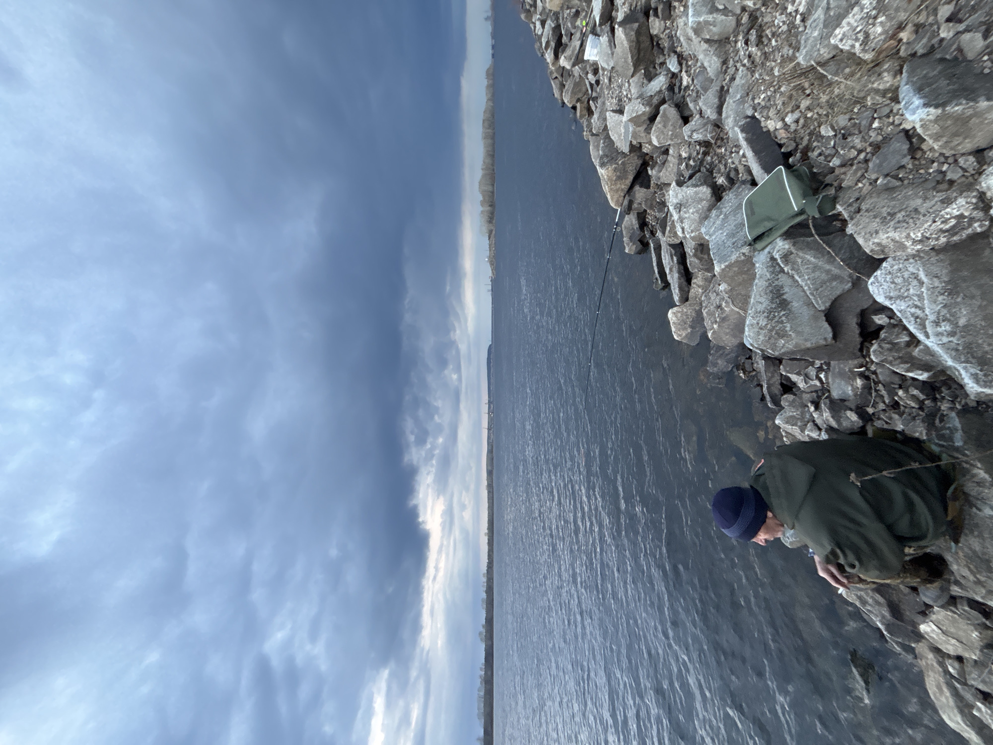 The image captures a scene on the shore of a river or large body of water, likely the Kama River based on the location of Chaikovsky, Russia. A man is positioned at the edge of the rocky bank, appearing to be focused on something near the water. He is wearing a dark blue knit cap and a dark green jacket. A fishing rod is visible extending out over the water. A small green bag is situated on the rocks nearby. The water is dark, reflecting the sky. The sky is overcast with heavy clouds. On the opposite shore, a strip of land is visible.