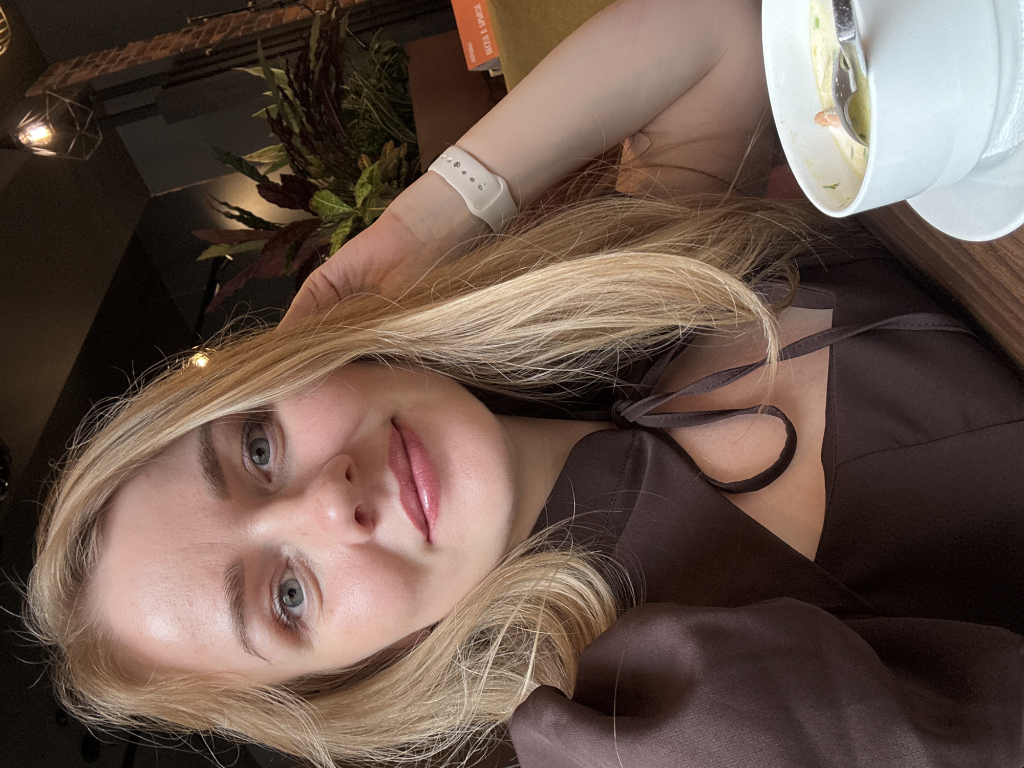 A young woman with blonde hair is posing for a selfie in what appears to be a restaurant setting. Her hair is swept to one side, and she is wearing a brown top with thin straps. A white smartwatch is visible on her wrist. Next to her is a bowl of soup on a wooden table. The background shows indoor plants and a decorative light fixture, suggesting a modern interior design.
There is no indication in the image that this is Saint Petersburg, Russia.