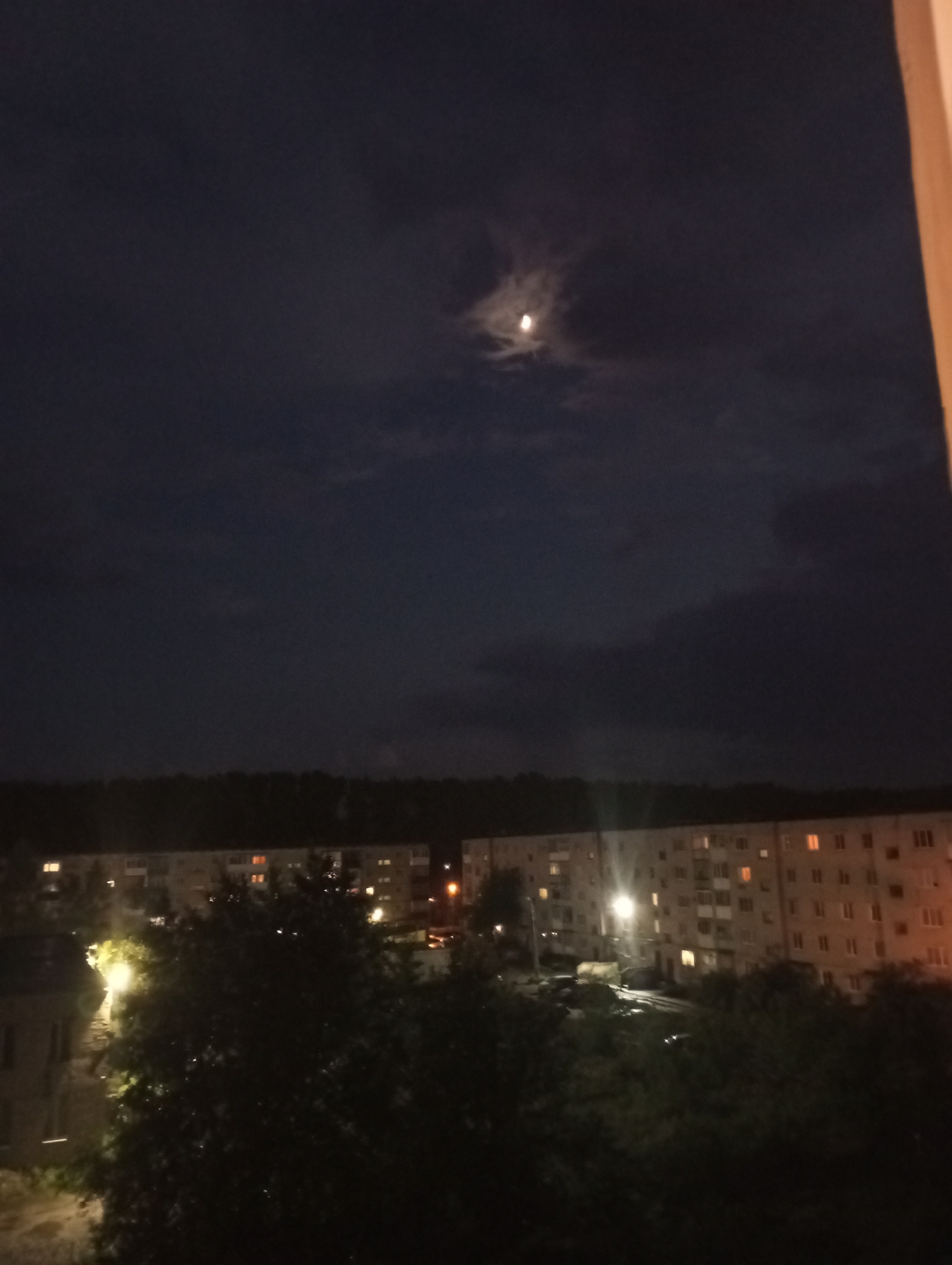 The image shows a night scene in what appears to be Kamensk-Uralsky, Russia. The sky is dark with clouds, partially obscuring the moon. In the distance, there are residential buildings with lit windows, indicating occupancy. Trees are visible in the foreground, their dark shapes contrasting with the illuminated buildings. The overall scene suggests a quiet, urban night.
