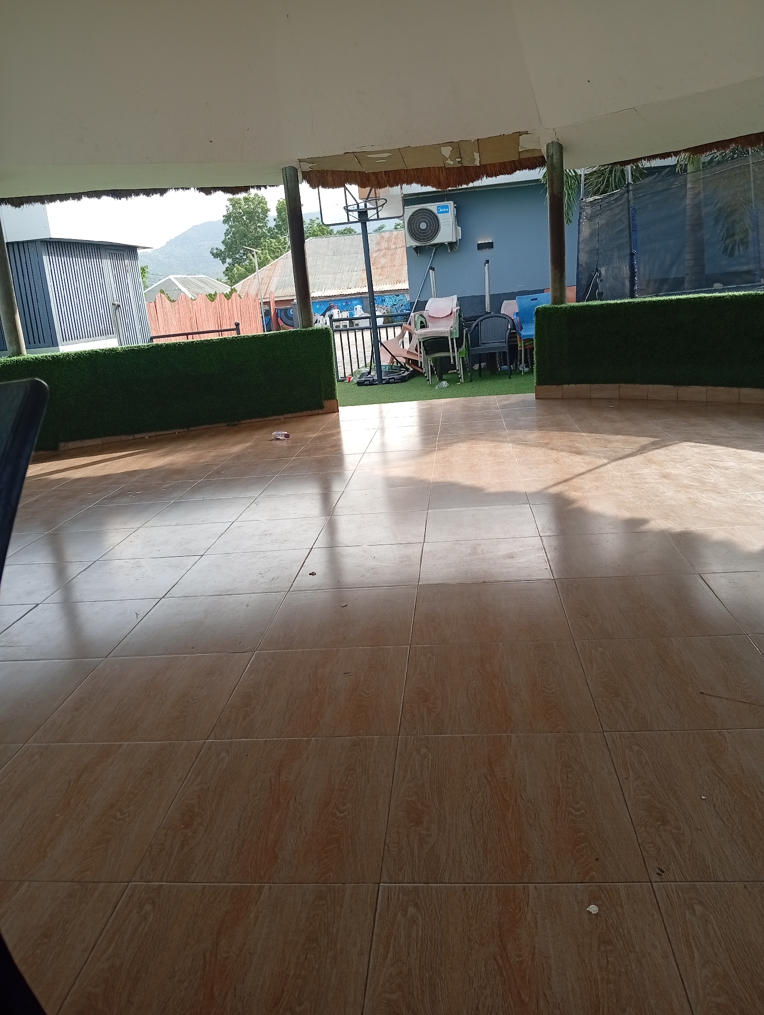 The image depicts a tiled patio or veranda in Adankolo, Nigeria. Artificial green hedges border the space. Various chairs and a basketball hoop are visible in the background. A building with an air conditioning unit is also present. The foreground consists mainly of brown, square tiles arranged in a grid pattern. Sunlight and shadows play across the tiled surface. No people are directly visible in the image.