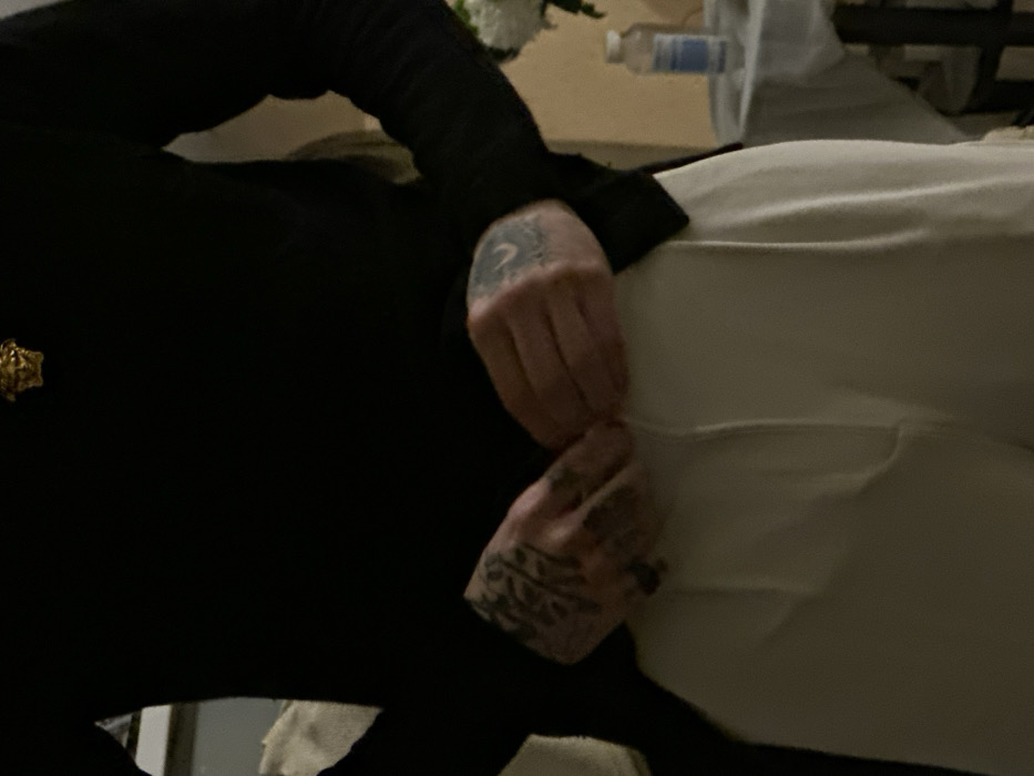 A person in a black, long-sleeved shirt and light-colored pants is visible. The individual has tattoos on both hands, including a moon design on one. Their hands are resting on the pants. A gold pin or badge is visible on the person's left side of the shirt. In the background, there is a water bottle, a white bag or cloth, and furniture. The lighting is dim, and the image is slightly rotated.