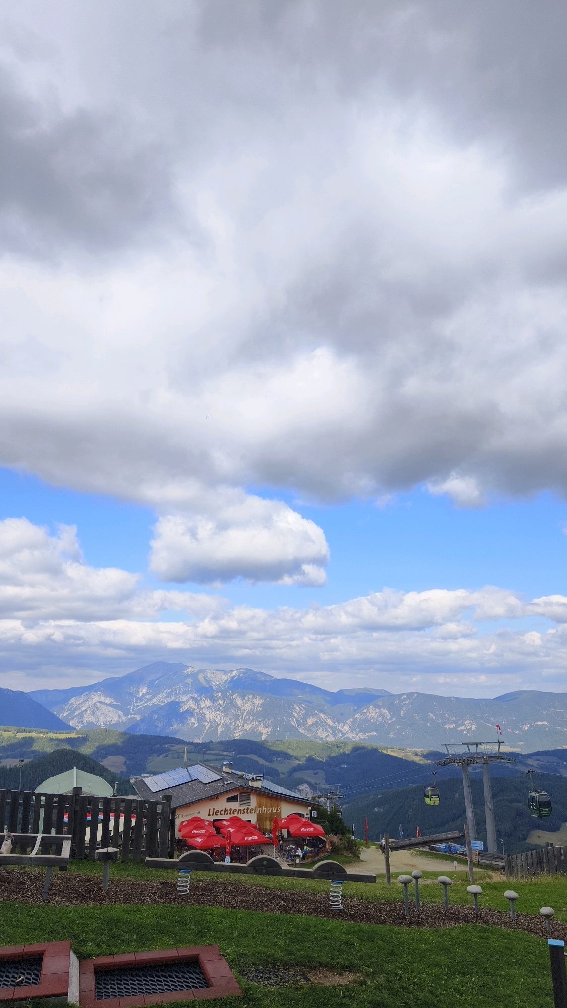 The image captures a scenic view from a mountaintop, likely in Semmering, Austria. In the foreground, a children's playground area is visible with a spring rider and a red and black brick structure. Beyond the playground, a building with solar panels on its roof is prominently featured, identified by the signage "Liechtensteinhau." Red umbrellas, associated with outdoor seating for the establishment, are open, and several people are seated at tables beneath them. To the right of the building, a ski lift with two gondolas can be seen ascending the mountain. The middle ground displays rolling green hills dotted with trees and other buildings. In the background, majestic mountains with rocky, exposed slopes rise under a dramatic sky filled with large, fluffy white and gray clouds. The sky transitions from a bright blue to overcast, suggesting varied weather conditions. The overall scene depicts a tourist destination offering both recreational activities and stunning natural landscapes.