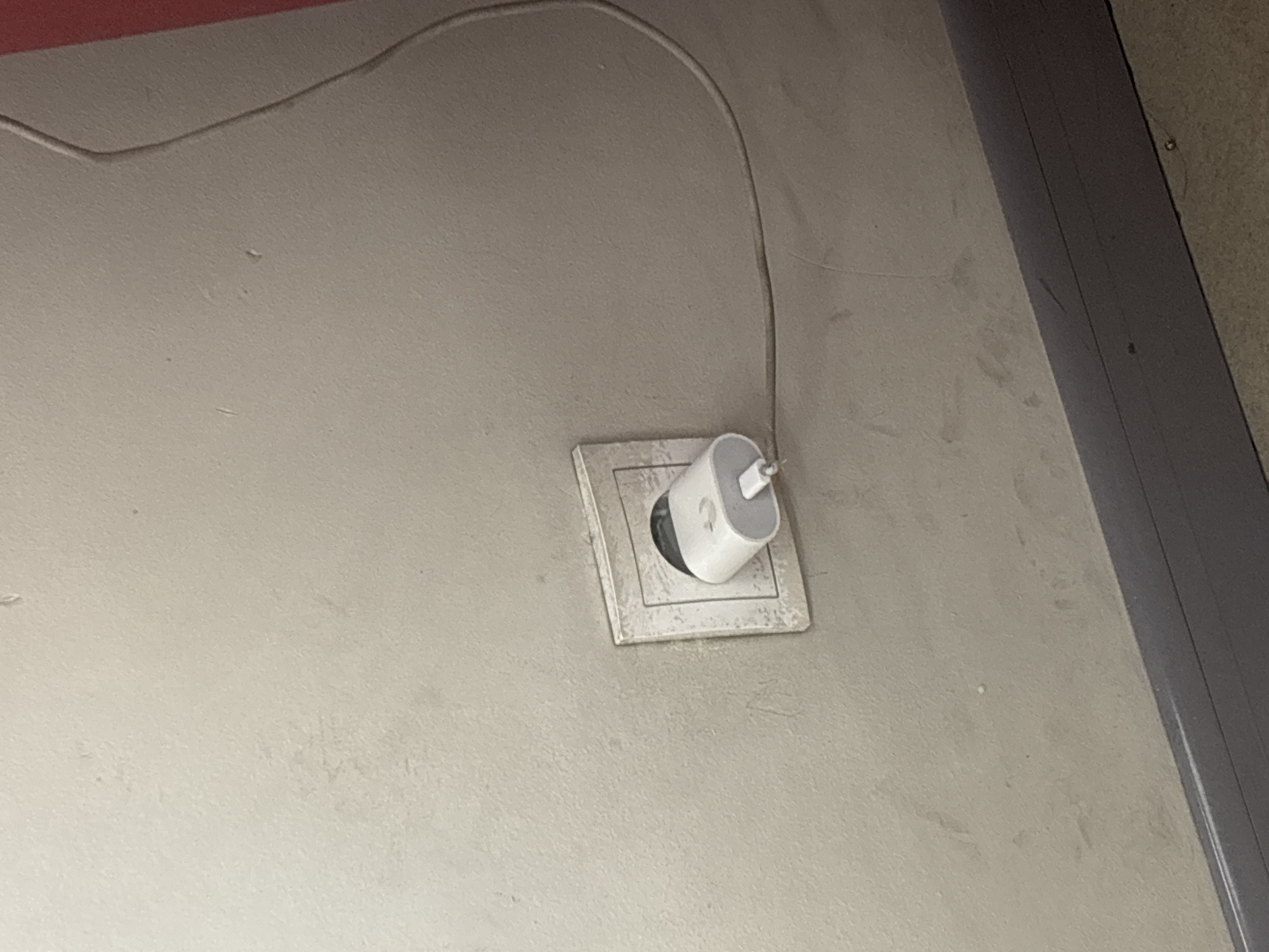 The image presents an indoor scene focused on a white electrical outlet and a charger adapter plugged into it, set against a light grey or beige textured wall. The outlet is a European-style two-pin socket, typical of the region, including Yerevan, Armenia.
A white, cylindrical charger adapter is inserted into the outlet at a slight upward angle. A thin, white cable extends from the top of the charger, curving across the upper portion of the wall towards the left, eventually disappearing out of frame. Faint, unidentifiable text or a logo is partially visible on the side of the charger.
The wall shows signs of wear and tear, with some dirt, smudges, and discoloration visible, especially around the outlet. In the upper left corner, a fragment of a red surface or object is present. To the right of the frame, a dark grey vertical structure, possibly a door frame or trim, runs along the wall. No specific activity is depicted, and the lighting suggests an interior environment.