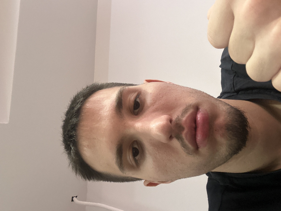 The image displays an eye-level, close-up self-portrait of an adult male indoors in Varna, Bulgaria. The subject has short, dark hair, brown eyes, a light skin tone, and a goatee. He wears a dark-colored t-shirt. His left hand is visible on the right side of the frame, clenched into a fist. The background consists of a plain white wall and ceiling, with a white cable or tube visible in the upper left corner. The male is looking directly at the camera.
