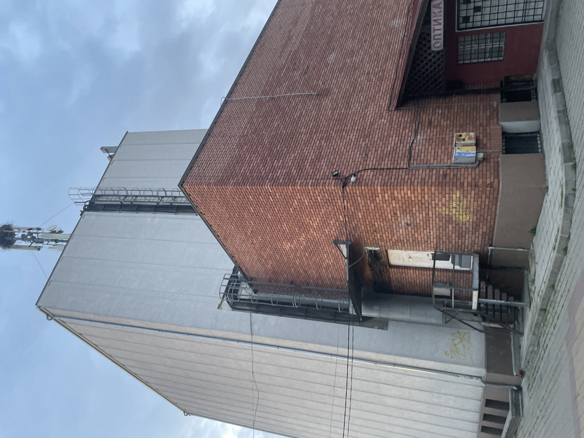 The image displays an urban scene in Ihtiman, Bulgaria, captured from a low-angle perspective looking upwards at a complex of buildings against an overcast sky.
On the left, a tall, light-colored building with a smooth, panelled facade extends towards the top of the frame. Near its upper edge, a metal access ladder or walkway with protective railings is attached. Atop this building, a telecommunication mast or cell tower equipped with multiple antennae is visible, and a large bird's nest is conspicuously built into its structure. Faint, dark-colored graffiti marks the lower section of this building's wall.
To the right and slightly in front of the light-colored building, a prominent corner of a reddish-brown brick building is featured. Its facade shows variations in brick color, with some areas appearing discolored or having faded yellow graffiti. Several electrical conduits and wires run along this brick wall, alongside a small security light and a yellow and blue electrical junction box.
Behind the brick building, a section of a darker red structure with a grid-patterned window is visible. A vertical sign on this red section displays the Cyrillic word "ОПТИКА" (Optika), which translates to "Optics" or "Optician's". Various grey pipes, ventilation ducts, and additional metal support structures are nestled in the narrow spaces between the buildings.
The sky is cloudy and grey, suggesting an overcast daytime. No people are visible, and no specific activities are taking place, with the focus solely on the architectural and utility details of the buildings. A paved surface, indicative of a sidewalk or street, is partially seen on the right side of the original image, bordering the structures.