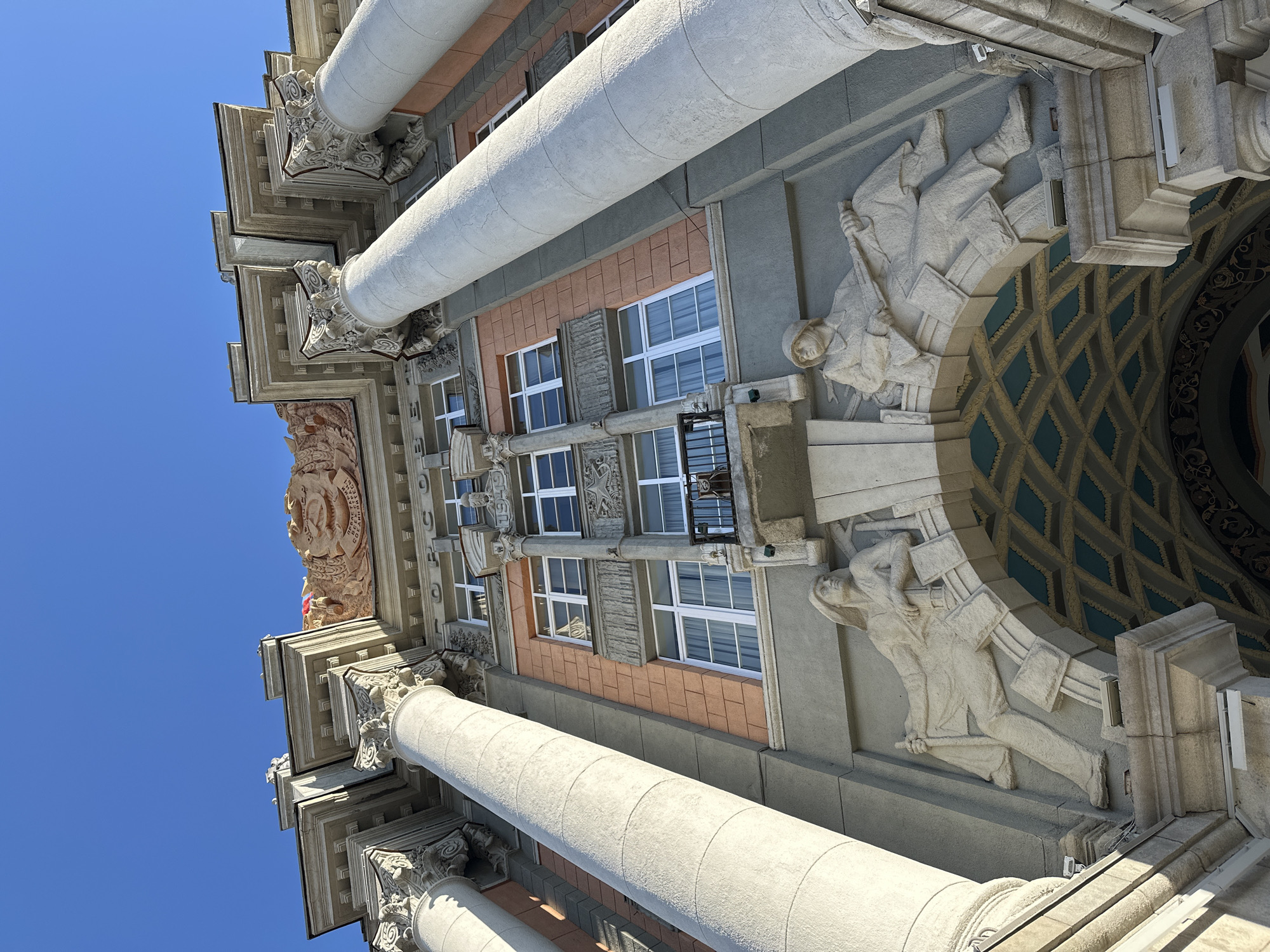 The image presents an upward-angled view of a building's facade in Yekaterinburg, Russia. The architectural style is characterized by neoclassical elements and socialist realism details. Massive white columns with ornate Corinthian capitals are prominent features. The facade is decorated with bas-relief sculptures of Soviet-era figures holding tools and books, indicative of workers and intellectuals. The building has multiple windows with white frames and a small balcony with a black railing. There are peach-colored brick elements as well as light grey stucco and a mosaic patterned interior visible behind the sculptures. The sky is clear blue.