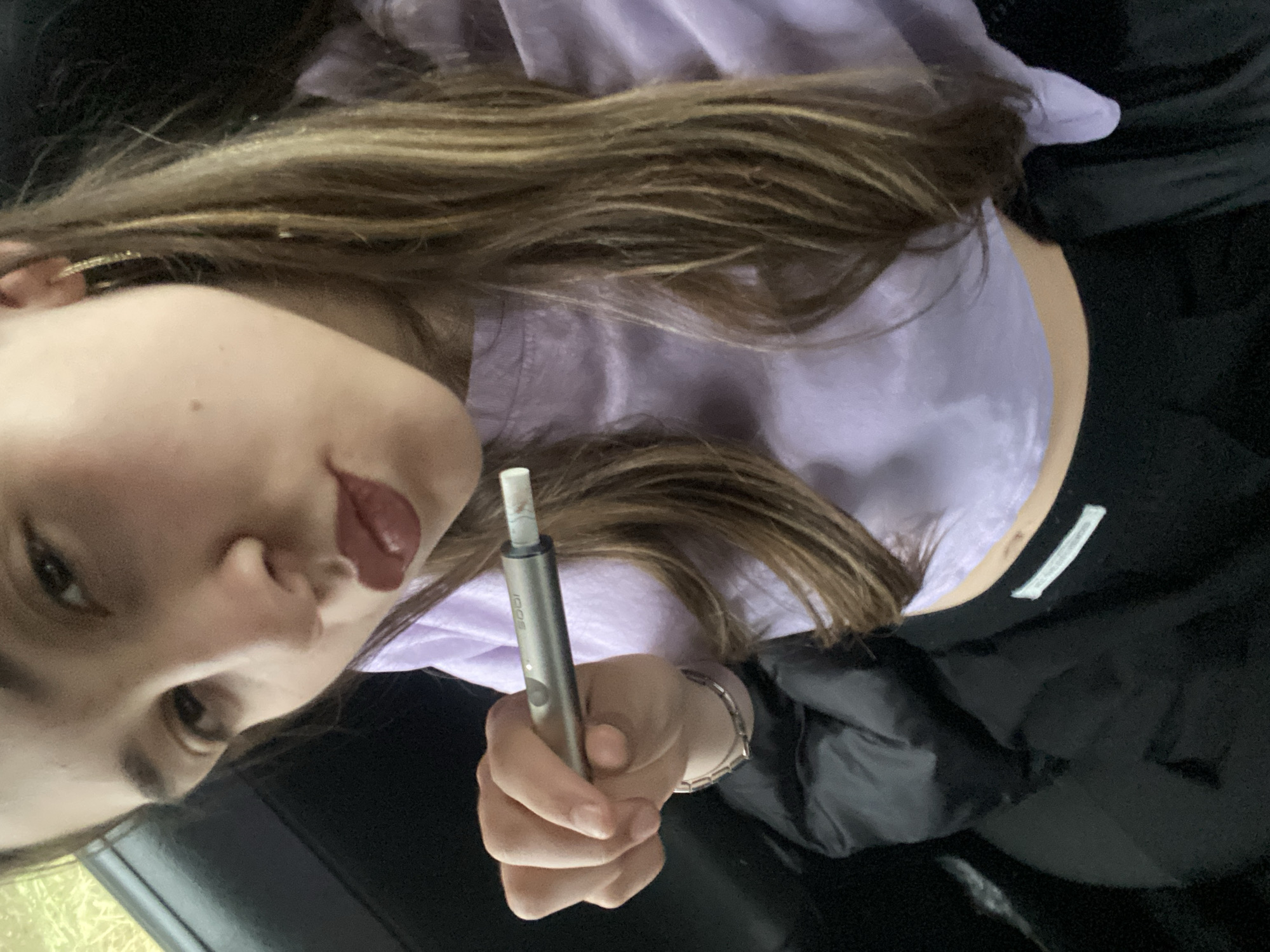 The image presents a selfie of a young woman, likely in her late teens or early twenties. She is holding a silver, pen-like device, possibly an electronic cigarette or similar vaping device. She is wearing a light purple cropped top that reveals a portion of her midriff. The environment appears to be indoors, possibly a car, suggested by the dark interior and glimpses of what could be an exterior through a window on the left side of the frame. Location details are not visually present and therefore cannot be determined.