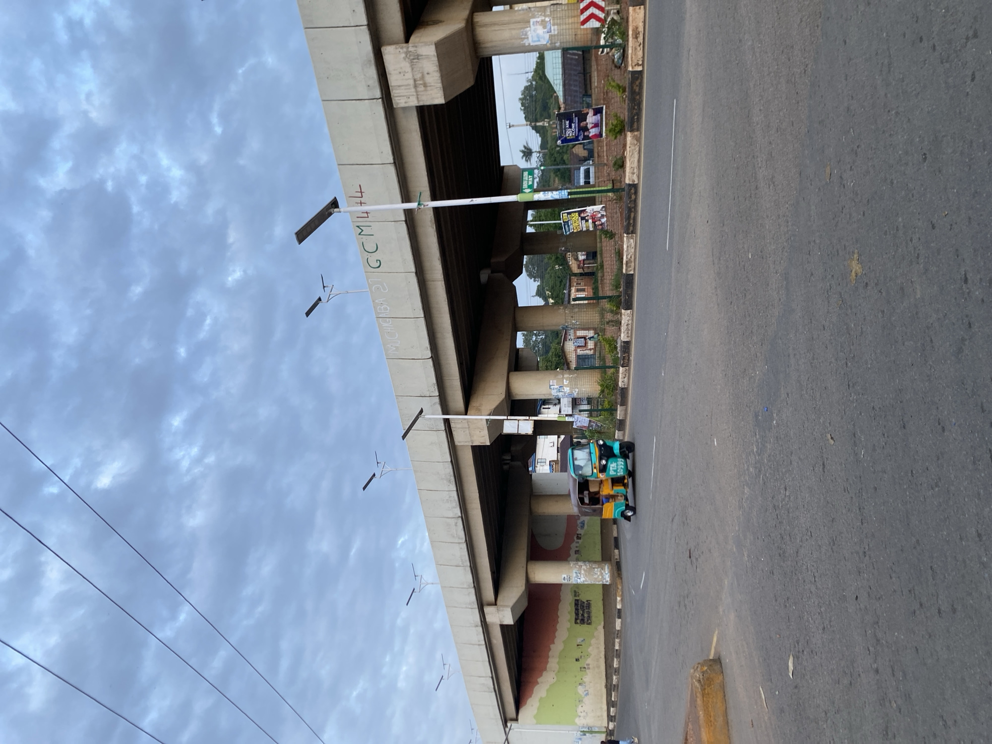 This image captures an overpass in Jos, Nigeria. A colorful auto-rickshaw, identified by the license plate "PTA 10399", is visible on the road beneath the overpass. The overpass itself is supported by large concrete pillars. Some of the pillars have graffiti and posters attached to them. In the background, there are buildings and lush greenery. The sky is overcast with scattered clouds. The scene appears to be a typical street view in Jos.