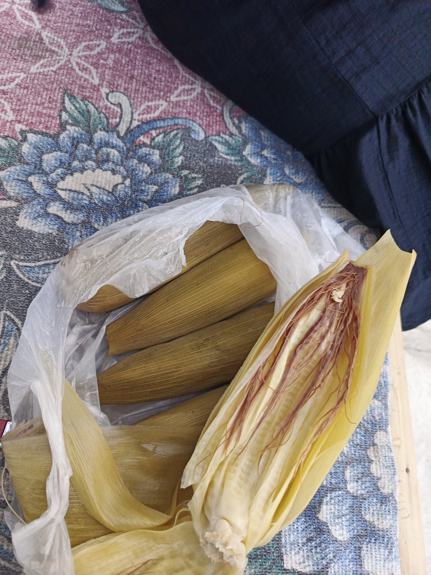 In Sumbersari, Indonesia, a bundle of corn husks, possibly containing tamales, rests in a clear plastic bag on a patterned fabric surface. One husk is peeled back, revealing the corn silk and a hint of the filling. The fabric has a floral design in pink, blue, and white. A dark blue fabric, likely clothing, is visible in the upper right corner. The lighting suggests an outdoor or well-lit indoor setting.