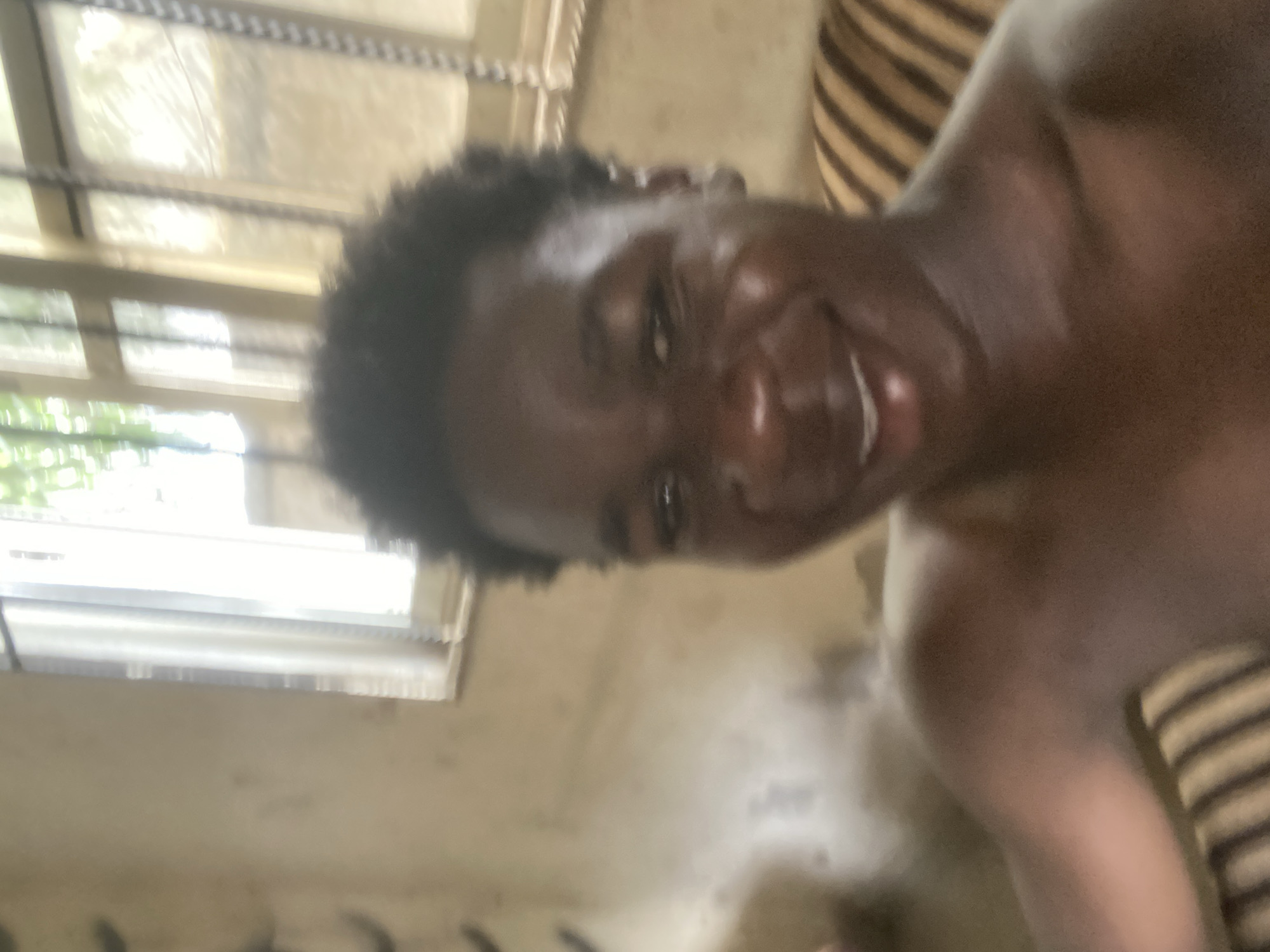 The image depicts a dark-skinned person with short, dark hair smiling. The person is shirtless and appears to be indoors. A barred window is visible in the background, suggesting a residential setting. The wall appears to be a pale color. The overall scene is blurry and suggests a snapshot taken in low-light conditions. No specific location can be definitively determined from this image. The statement about Mararaba, Nigeria is conjecture.