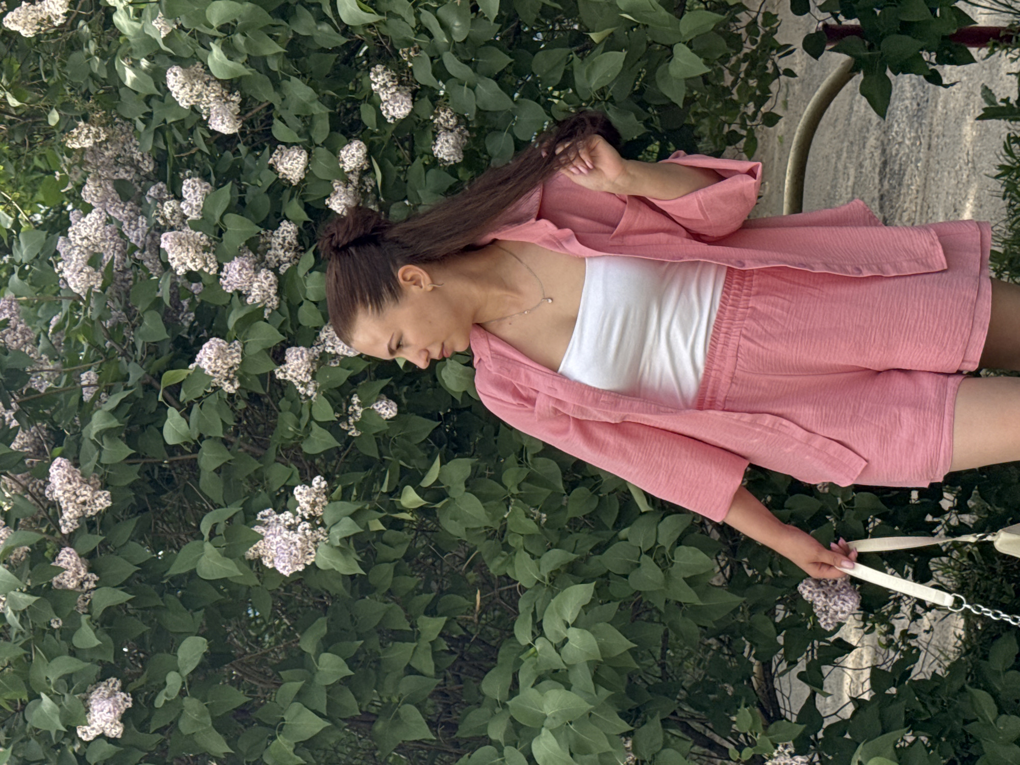 A young woman stands next to a blooming lilac bush. She has brown hair pulled back into a bun and is wearing a pink outfit with a white top. She holds a white purse with a chain strap in her right hand, and her left hand is touching her hair. The location appears to be an outdoor area in Chaikovsky, Russia, based on prior knowledge. The bush is full of flowers and green leaves.