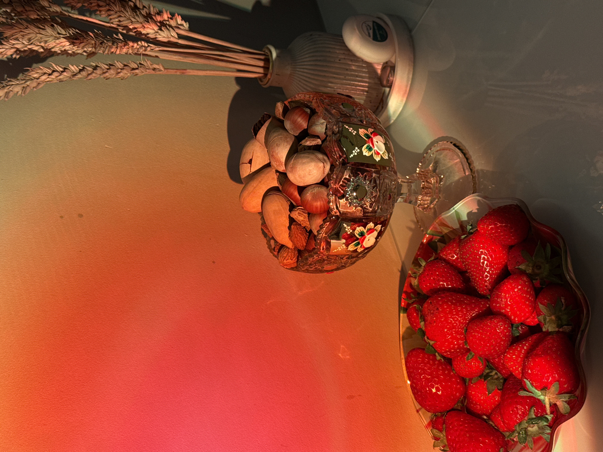 A high-angle studio shot features a still life composition. In the upper left, a vase filled with dried wheat stalks casts a shadow. Next to it, a decorative glass bowl containing mixed nuts sits atop a crystal bowl with a strawberry motif and a base filled with strawberries. The backdrop is a gradient of orange-red on the left and blue-grey on the right.
