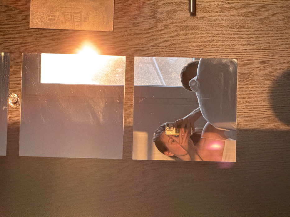A woman and a man are reflected in a square mirror on a wood-paneled wall. The woman holds a smartphone up towards the mirror. A second mirror reflects sunlight. The man stands behind the woman, out of focus.