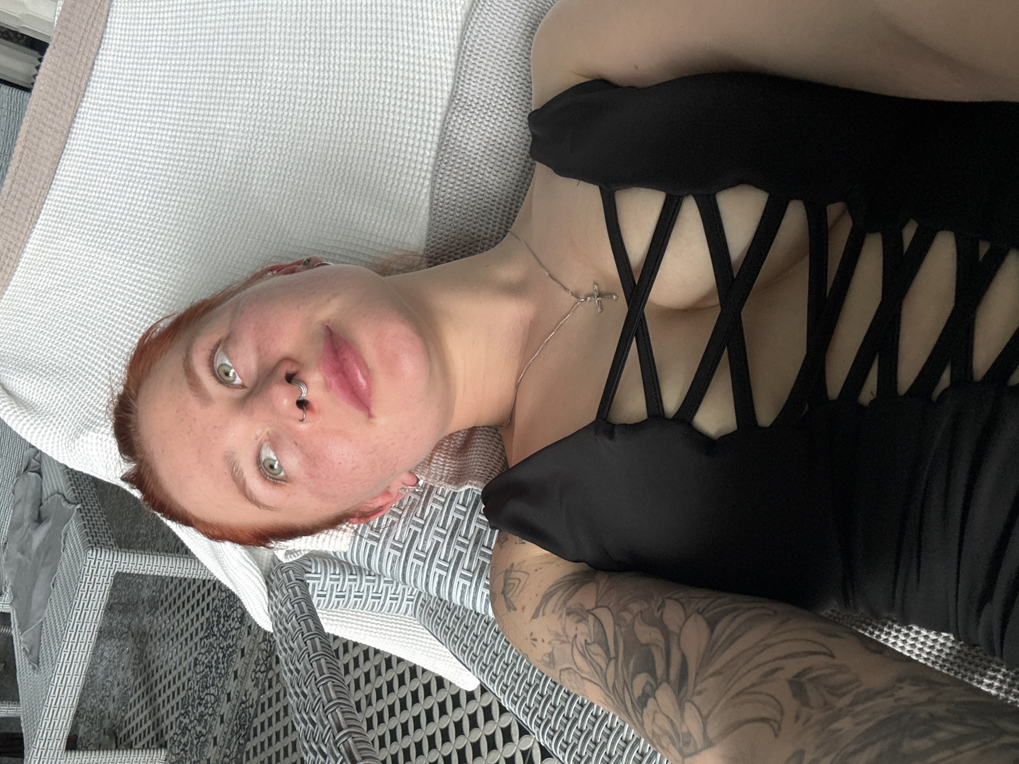 A woman with light skin, red hair, and a nose ring is lying on a textured white and grey cushion. She is wearing a black top with a crisscross design across the chest, and a silver necklace with a cross pendant is visible on her neck. Her left arm, covered in a large black and grey tattoo, is resting on the cushion. The cushion rests on a white and grey woven chair. The image is taken at an angle, with the camera positioned above the woman. It's impossible to tell if this is Rostov-on-Don without more context.