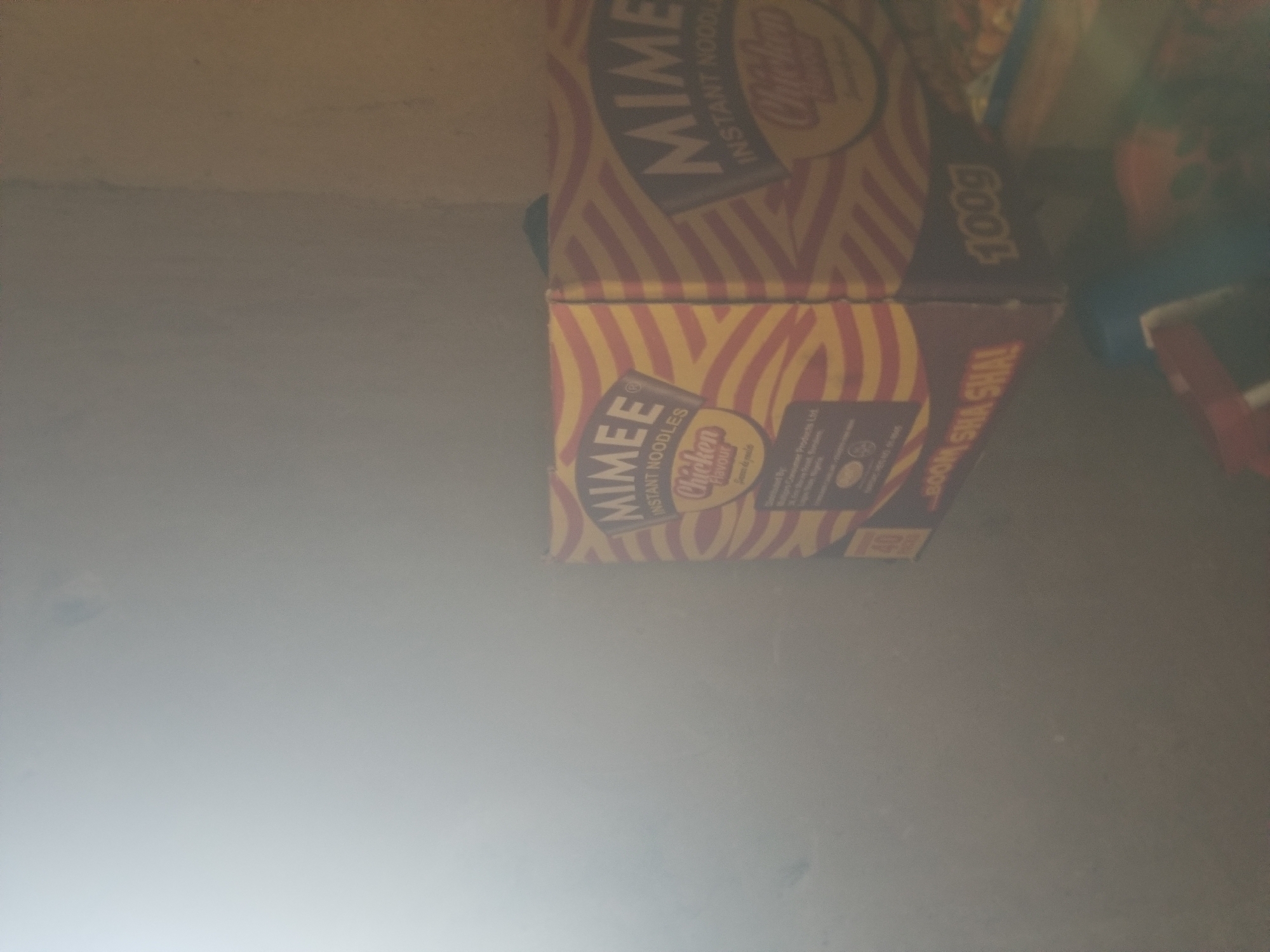 The image shows a corner of a room with a bright light source on the left side of the frame. In the center right, there are two boxes of instant noodles, branded "Mimee" with a "Chicken Flavour" label. The boxes have a red and yellow striped pattern. One box is partially visible on the right and displays "100g". The box on the left is more visible and shows "40 packs" and "BOOM SHA SHAL". The environment appears to be dusty, with a wall visible in the background. The location of Jauro Yinu, Nigeria, is mentioned in the prompt but there are no visual cues in the image to confirm or suggest this specific location.