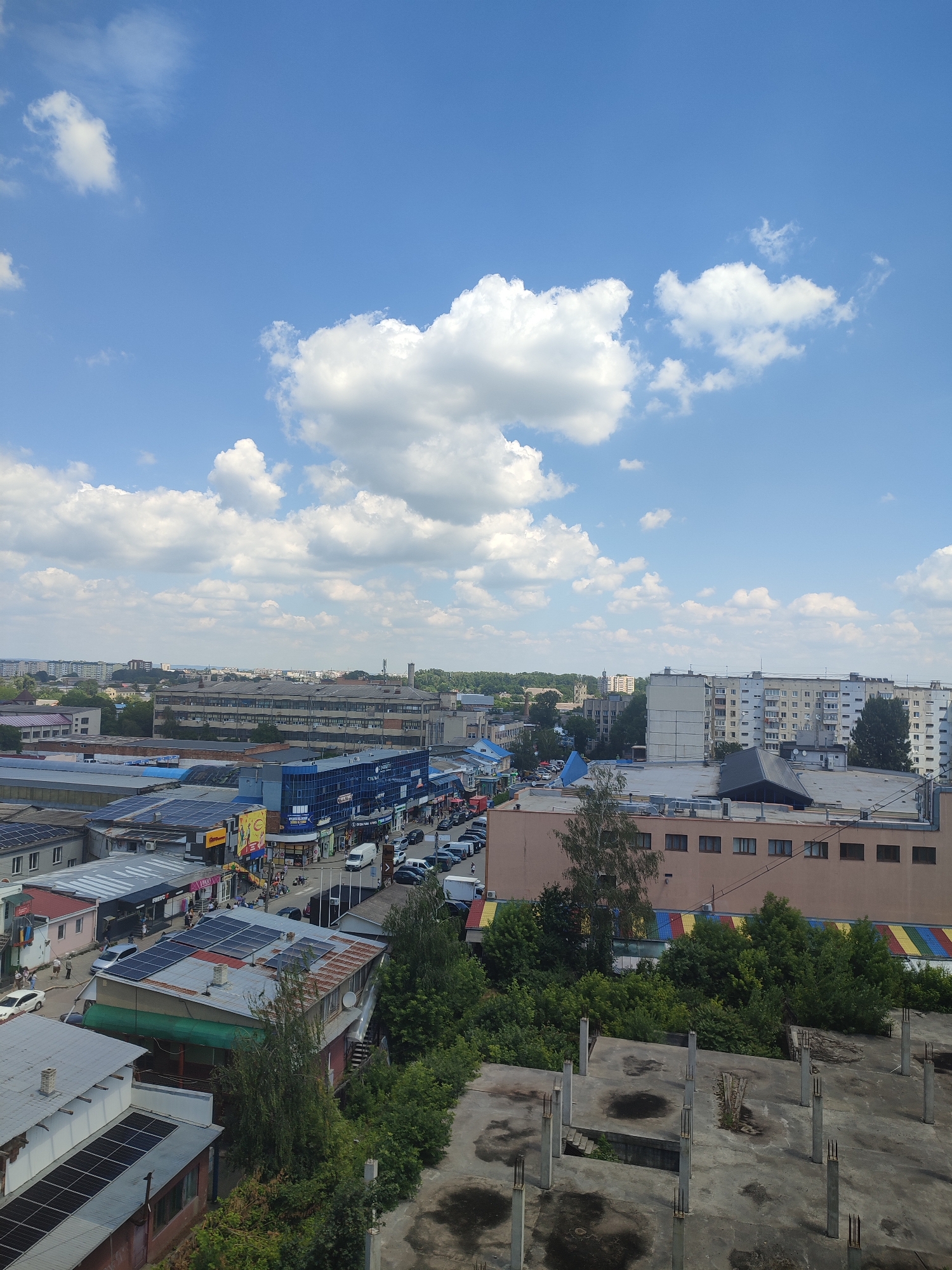 An aerial view captures a bustling urban scene in Chernivtsi, Ukraine, under a bright blue sky dotted with fluffy white clouds. The image showcases a dense urban landscape with a variety of buildings, ranging from commercial establishments with colorful awnings to residential blocks and industrial-looking structures. Several roads are visible, lined with parked cars and a few moving vehicles, indicating active traffic.
In the foreground, a partially constructed building with visible concrete pillars and rebar suggests ongoing construction or an abandoned project. The roofs of many buildings are adorned with solar panels, highlighting an effort towards renewable energy. Green trees and foliage are interspersed throughout the urban environment, softening the otherwise hard-edged cityscape. The presence of people walking on sidewalks and near shops indicates daily life and commercial activity. The overall impression is of a dynamic and developed city with a blend of modern infrastructure and everyday urban life.