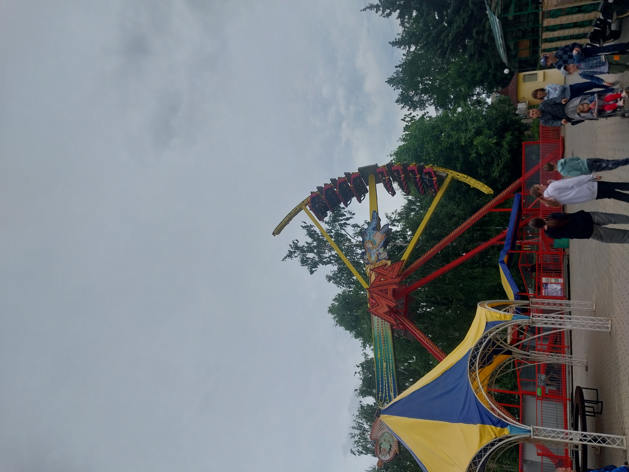 The image shows an amusement park ride in Orenburg, Russia. The ride is a large, pendulum-style attraction with multiple seats occupied by people. It is painted in bright colors like red, yellow, and blue. Below the ride, there is a yellow and blue tent-like structure. The sky is overcast with gray clouds. To the side, there is a group of people, some of whom are standing near a fence. Trees are visible in the background, adding to the park-like setting. The ground is paved.