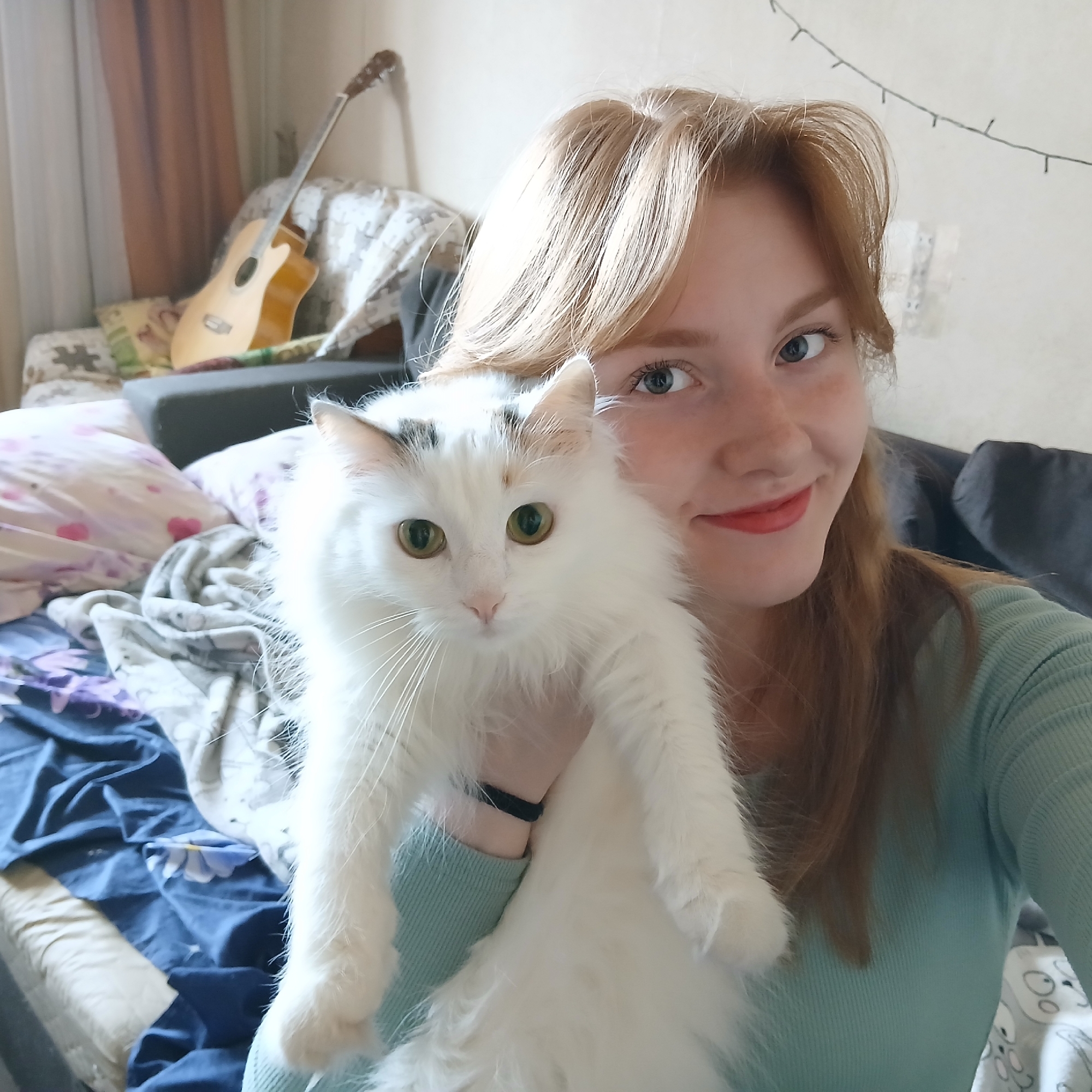 A Caucasian female with strawberry blonde hair is holding a white, long-haired cat with green eyes. The female has light skin, blue eyes, and is wearing red lipstick. She is wearing a light green ribbed long-sleeve shirt. The cat has some black markings on its head.
In the background, there is a bed with blue and white bedding and several pillows. Leaning against the wall behind the bed is an acoustic guitar. On the wall behind the female and the guitar, there is a string of lights. The location is inside a residence, likely in the city of Unknown, Unknown.