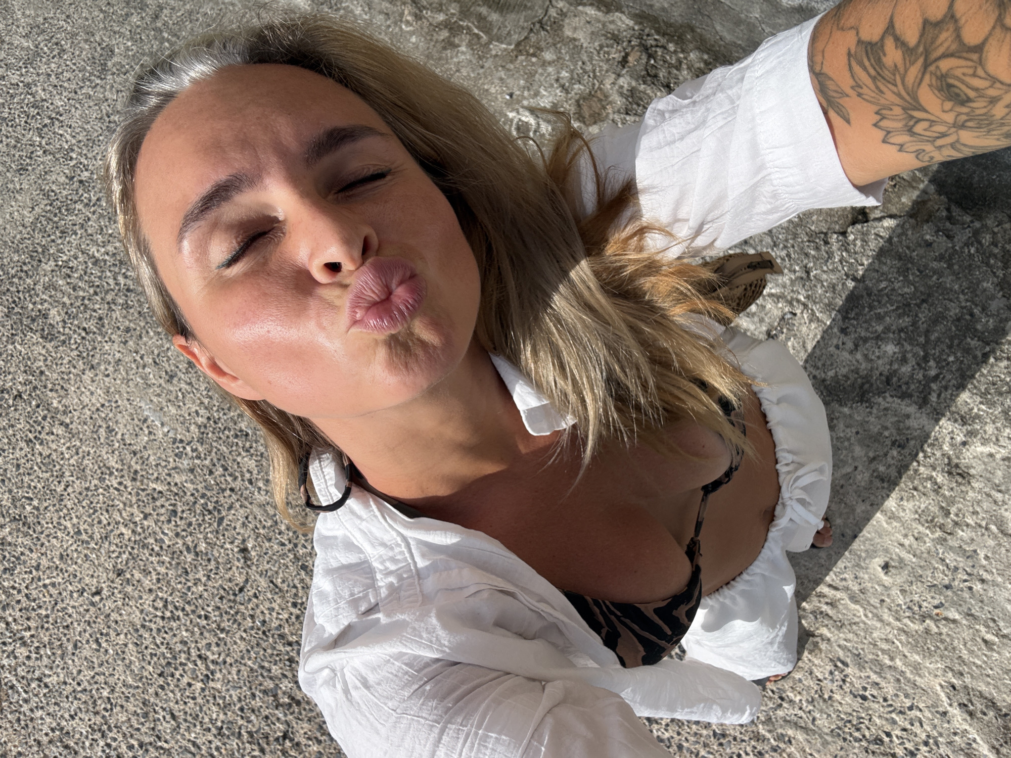 An overhead shot shows a blonde woman with her eyes closed and lips puckered in a kiss, taken outdoors on a textured grey surface. She is wearing a white button-up shirt, unbuttoned to reveal a patterned bikini top. Her right arm, visible on the right side of the frame, displays a tattoo of floral patterns. The scene is illuminated by sunlight, casting a shadow on the ground. The location is likely a public outdoor area in Ko Samui District, Thailand, given the textured ground which resembles a pathway or an unpaved area common in tropical climates.