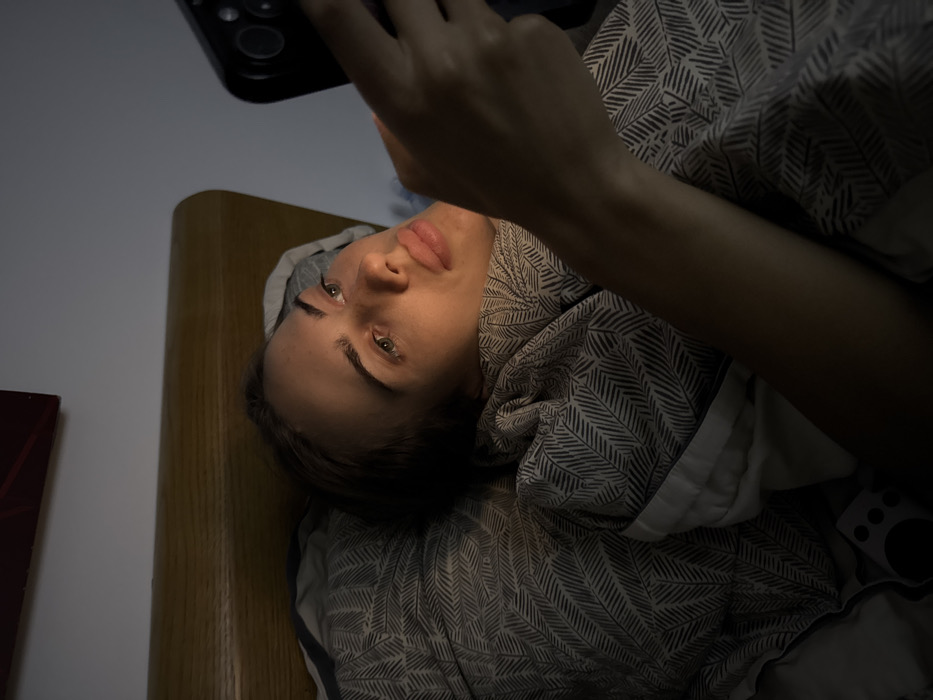 A person lies in bed, head turned towards the camera, holding a smartphone. Their face is softly illuminated by the phone's light, highlighting their eyes and lips. The bedding has a patterned design. The person's arm holding the phone is visible in the foreground. Part of a headboard is on the left, and a red object can be seen next to it. The room appears to be dimly lit.