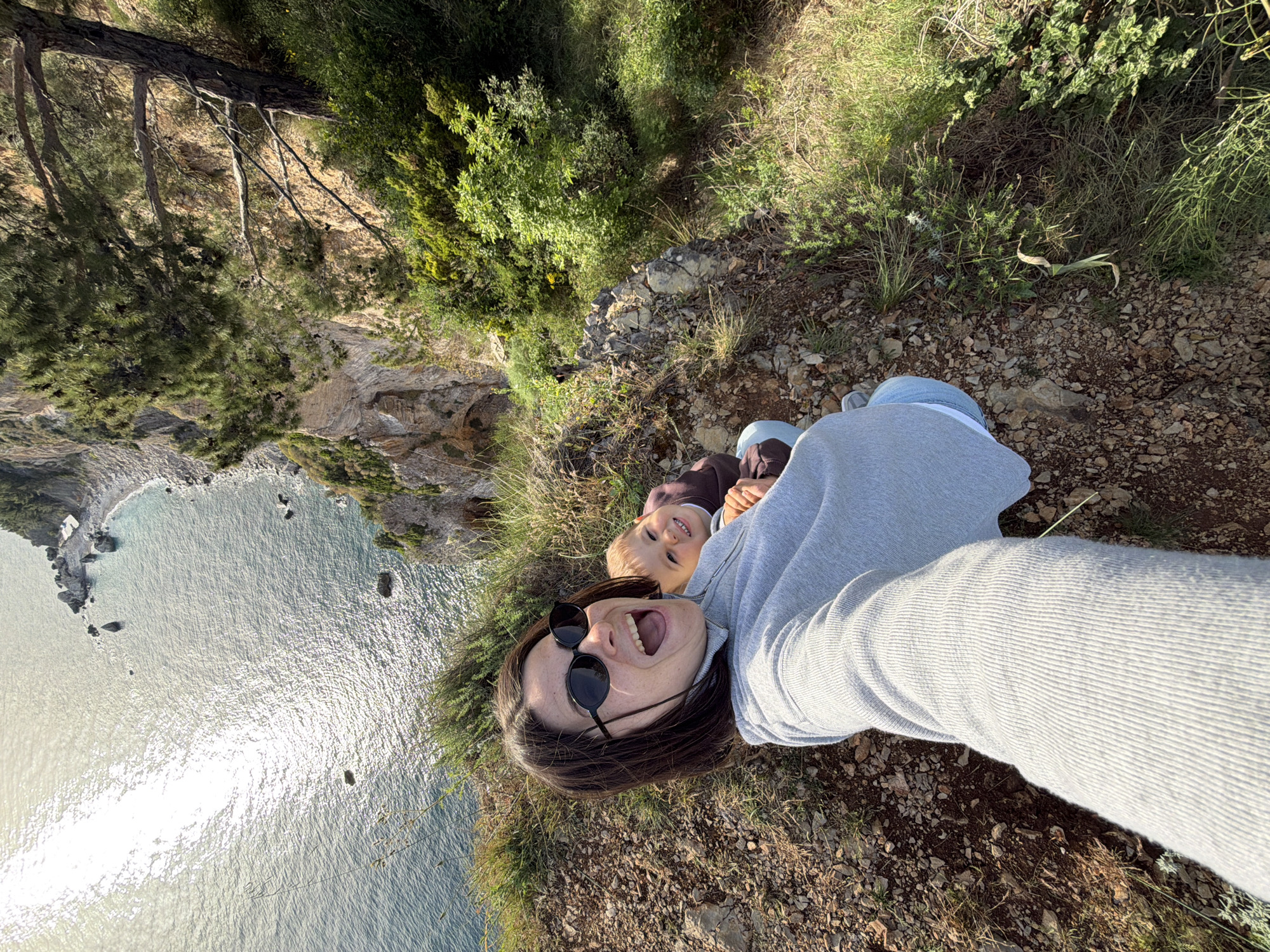 A selfie photograph captures a woman and child lying prone on a rocky hillside. The location is suggested to be near Budva, Montenegro, given the view of the Adriatic Sea and the coastal terrain. The woman is wearing dark sunglasses and a light grey sweater and is smiling with her mouth open. A young child is next to her, facing towards the camera. They are on terrain covered with small rocks, dry grasses, and sparse vegetation. In the background, there's a view of a bay or cove with clear blue water and rocky cliffs. A few scattered trees and bushes grow along the edges of the cliffs and the hillside.