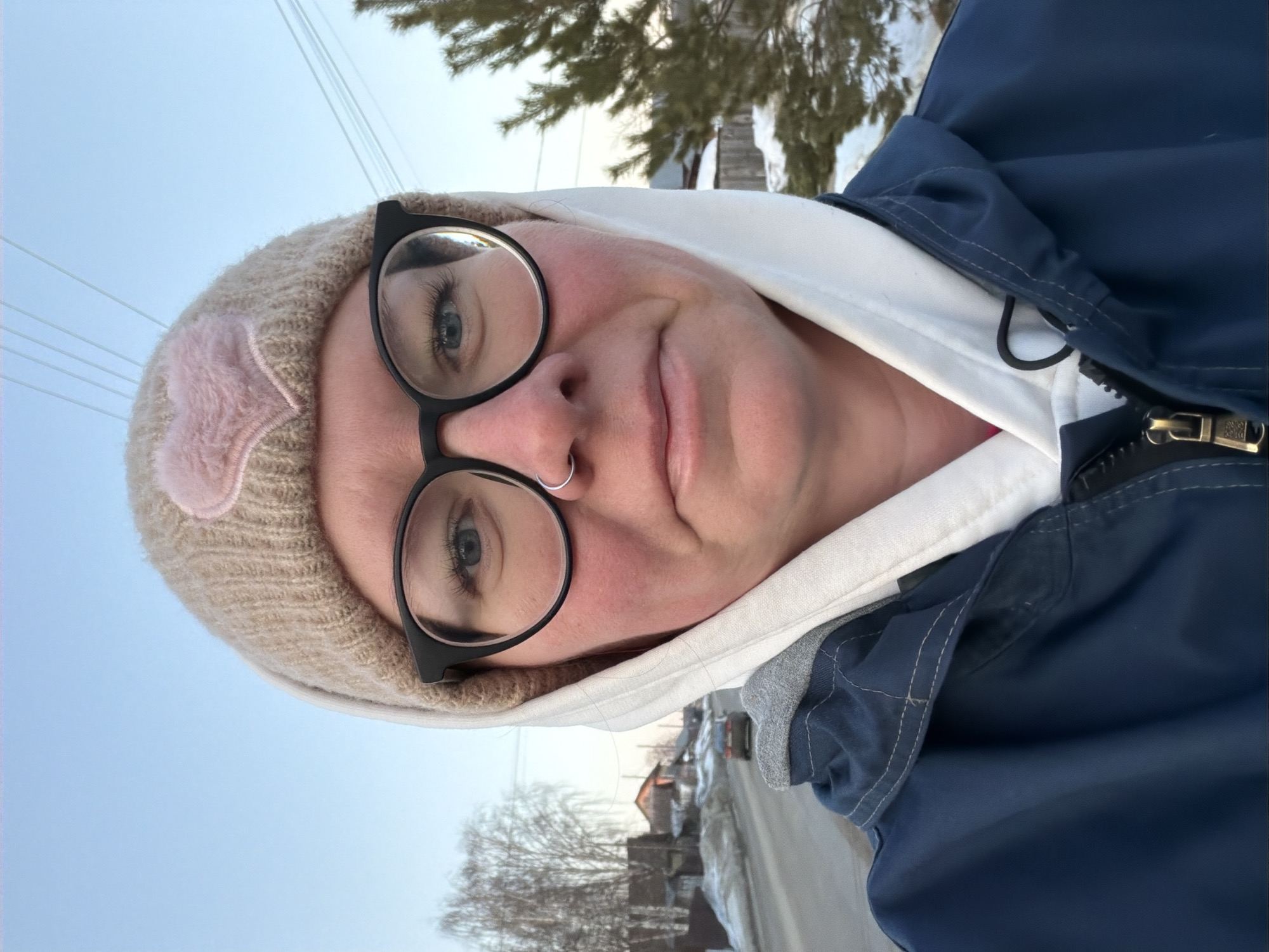 An outdoor portrait captures an adult female from the chest up, positioned against a winter residential street in Chaikovsky, Russia. The subject faces the camera directly with a neutral expression, wearing a beige knitted beanie adorned with a pink fuzzy heart, black-rimmed glasses, and a silver hoop nose ring in her left nostril. She is dressed in a white hooded sweatshirt with the hood up, beneath a dark blue zipped jacket. Her cheeks exhibit a slight flush. The background reveals a paved road, partially covered with snow and slush, extending into the distance. On the left, several detached houses and bare deciduous trees line the roadside, with a dark-colored sedan visible driving away from the viewer. On the right, a coniferous bush and sections of a snow-covered fence are present. Overhead power lines traverse the clear, light blue sky.