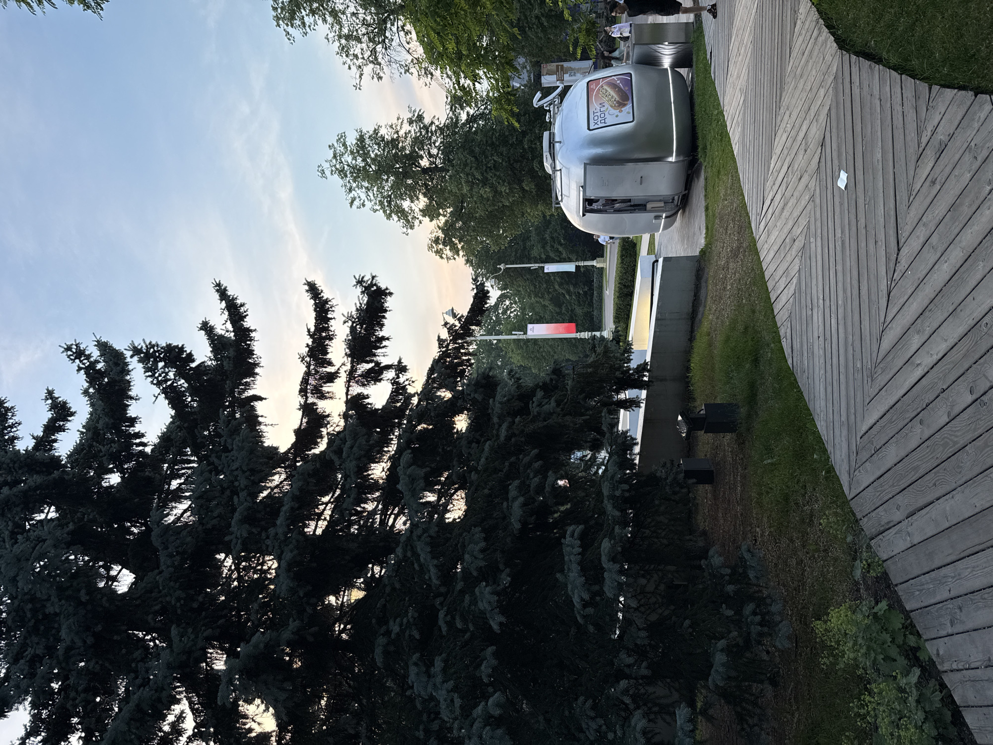 The image shows an outdoor scene in Khimki, Russia, possibly near a park or public space. In the foreground, a curved wooden walkway dominates the lower portion of the frame, its planks laid in a diagonal pattern. To the left, a large, dark green coniferous tree with dense foliage is prominent, its branches extending over the walkway.
Further into the scene, a silver, teardrop-shaped mobile food kiosk is parked on a paved area. A sign on the side of the kiosk displays an image of a hot dog and text in Russian that appears to read "ХОТ-ДОГИ" (Hot Dogs). A person is visible through the open service window of the kiosk.
Behind the kiosk and to the left, a concrete structure, possibly a low wall or planter, is visible. Beyond that, several lampposts and banners are spaced along a pathway. In the background, lush green trees form a dense line, and a pale sky with wispy clouds suggests it is either early morning or late evening.
Several people are scattered throughout the scene, walking or standing near the kiosk and the pathway. One person in the foreground on the right is wearing black shorts and a t-shirt and appears to be walking away from the camera. The overall environment suggests a public area designed for recreation and possibly commerce.