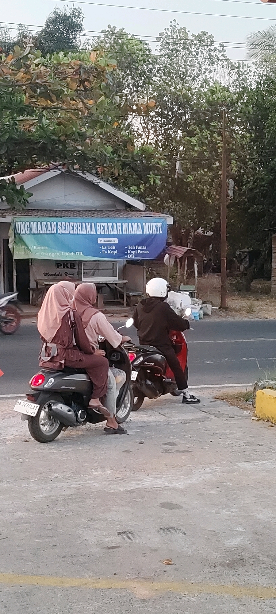 The image depicts a street scene in Bentok Kampung, Indonesia, with a focus on a roadside stall and two motorbikes. In the foreground, a dark gray scooter is stopped, carrying two individuals wearing headscarves. The passenger in the back is wearing a light brown headscarf and a maroon outfit, while the person in front wears a light pink headscarf and a matching outfit. They are positioned on the scooter's seat, facing away from the camera. Behind this scooter, a red motorbike is also stopped, with a person wearing a white helmet and a dark hooded jacket riding it.
A prominent blue banner is stretched across the front of a small, simple building. The banner reads "UNG MAKAN SEDERHANA BERKAH MAMA MUKTI" and lists various drinks such as "Es Teh," "Es Kopi," "Teh Panas," and "Kopi Panas." Below the banner, a table with items on it is visible. The building itself appears to be a modest structure with a white roof and walls.
The environment includes lush green trees and foliage in the background, suggesting a tropical or subtropical setting. A utility pole is also visible to the right of the frame. The road surface is paved, with a yellow line painted along the edge in the lower right portion of the image. The overall lighting suggests it is daytime, possibly late afternoon due to the warm tones. The scene is one of everyday life, with people utilizing motorbikes for transportation and patronizing local food and drink establishments.