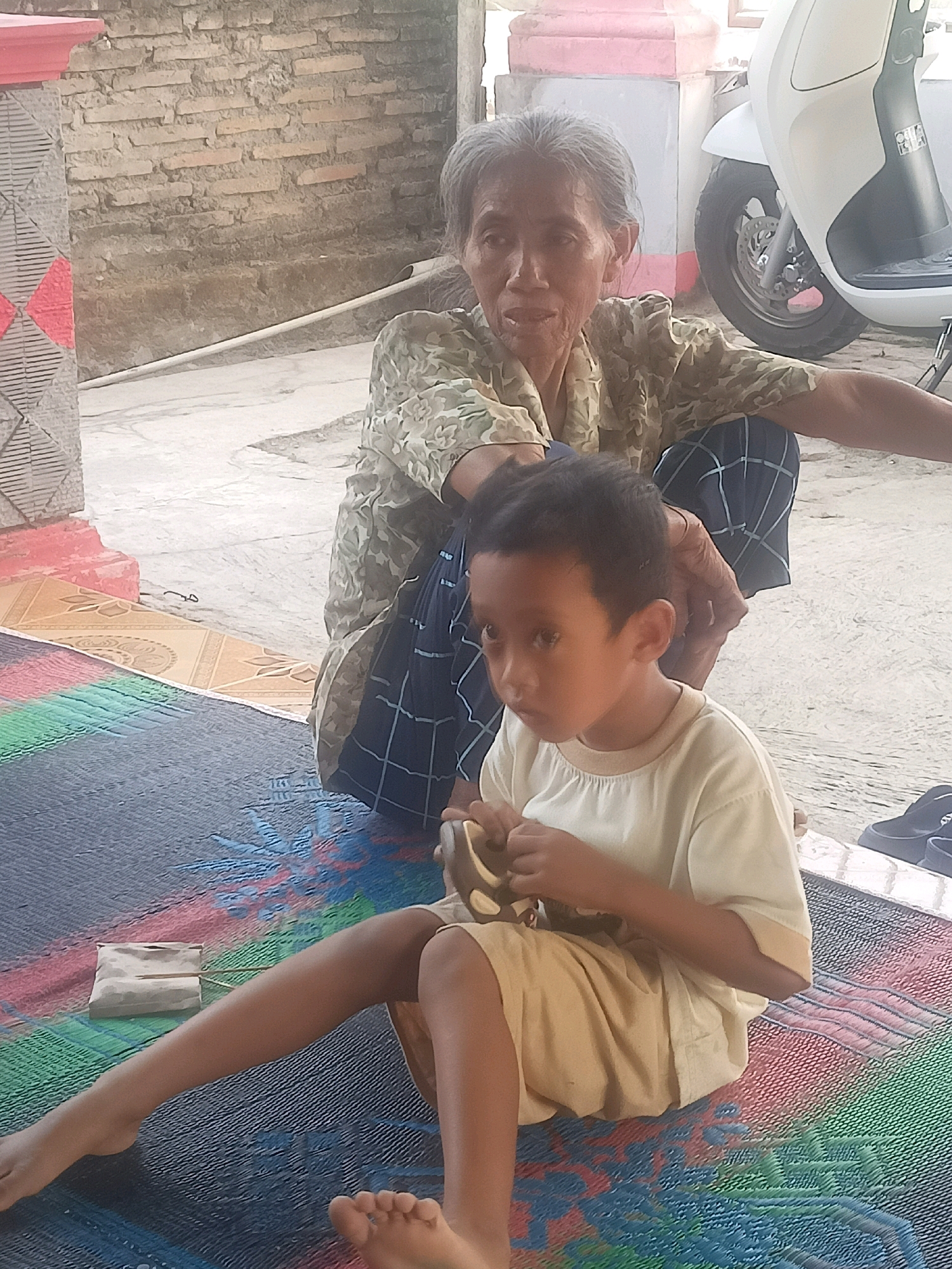 In Karangtalun, Indonesia, an outdoor scene depicts an elderly woman and a young boy sitting on a colorful woven mat. The woman, with gray hair and weathered skin, wears a patterned olive green blouse and dark blue checkered pants. She is partially behind the boy, her hands possibly tending to him. The boy, facing left and slightly away from the camera, has dark hair and is wearing a pale yellow t-shirt and light brown shorts. He is holding a brown object in his hands. In the background, a brick wall and a white scooter are visible, along with a pink and white structure. The ground is paved, and the mat they are sitting on features stripes of blue, green, red, and yellow.