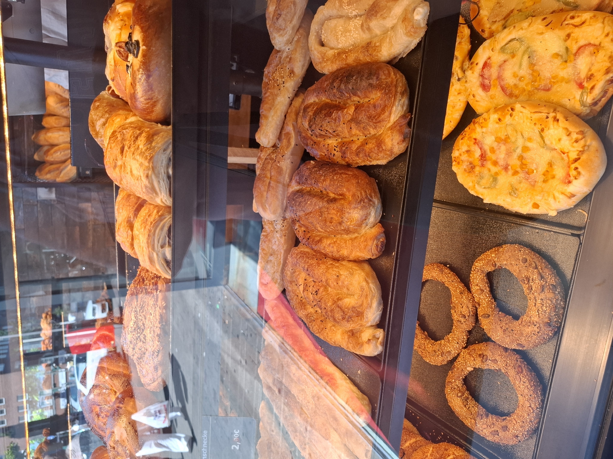 This is a photograph of a display case in a bakery, likely in Hamburg, Germany. The display case is filled with a variety of baked goods, including pastries, bread, and savory items. On the top shelf, there are large, golden-brown pastries that appear to be sweet or savory turnovers. Below this, on the second shelf, are several long, flat pastries and some circular pastries topped with sesame seeds and other grains. The third shelf features more substantial, golden-brown pastries, some with visible sesame seeds, and a swirled pastry. The bottom shelf contains three ring-shaped breads, possibly bagels or simits, covered in seeds, and several small, flatbreads topped with cheese and vegetables, resembling miniature pizzas or flatbreads. The background is slightly blurred, showing a reflection of a street scene with buildings and a vehicle. The lighting suggests an indoor environment, possibly a storefront. There are no discernible individuals in the foreground, but the reflection might show people or cars outside. The overall impression is a well-stocked bakery display.