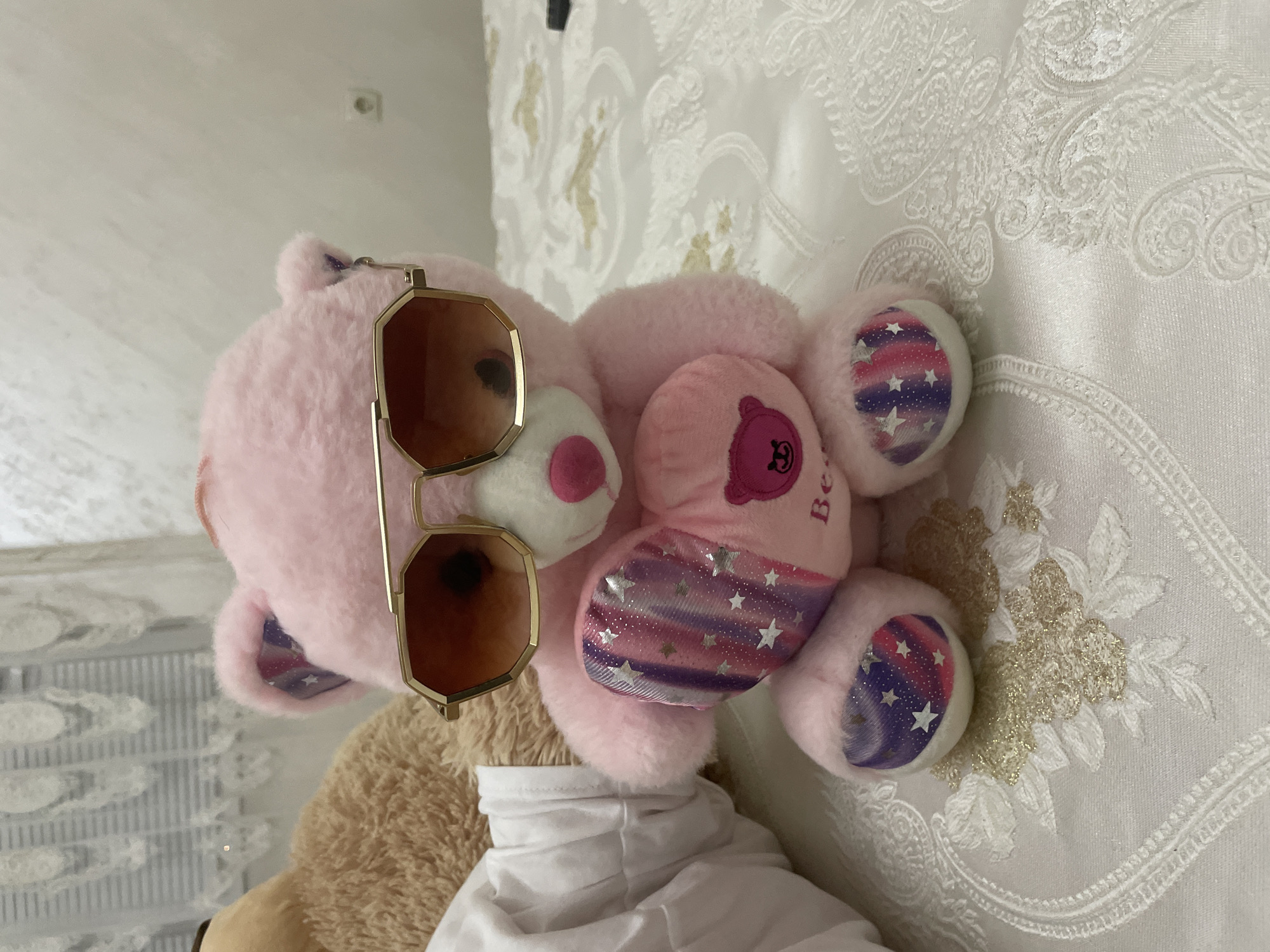 The image displays two teddy bears positioned on a decorative white bedspread indoors. The primary subject is a fluffy pink teddy bear, lying on its back, adorned with stylish gold-framed, hexagonal-shaped sunglasses with dark lenses. This pink bear has a white snout with a small pink nose, and its paws and feet are made of a different fabric featuring a sparkly, gradient pattern of purple, pink, and blue, speckled with white stars. On its left arm or paw, a darker pink patch shows a bear outline and the partially visible text "Bea".
To the lower left, part of a second teddy bear, appearing beige or light brown, is visible, wearing a white, high-collared shirt or top. Both teddy bears are resting on a white bedspread that features an intricate, possibly embroidered or jacquard, pattern of floral and paisley designs, with some accents of gold thread. In the background, a light-colored wall is visible, with a single electrical outlet positioned in the upper left corner of the frame.
The scene depicts a static, playful arrangement of the teddy bears. There is no specific activity taking place. The overall setting appears to be a domestic interior, likely a bedroom. The light suggests daytime, though no direct light source is visible. The European-style electrical outlet is consistent with the provided location context of Ihtiman, Bulgaria.