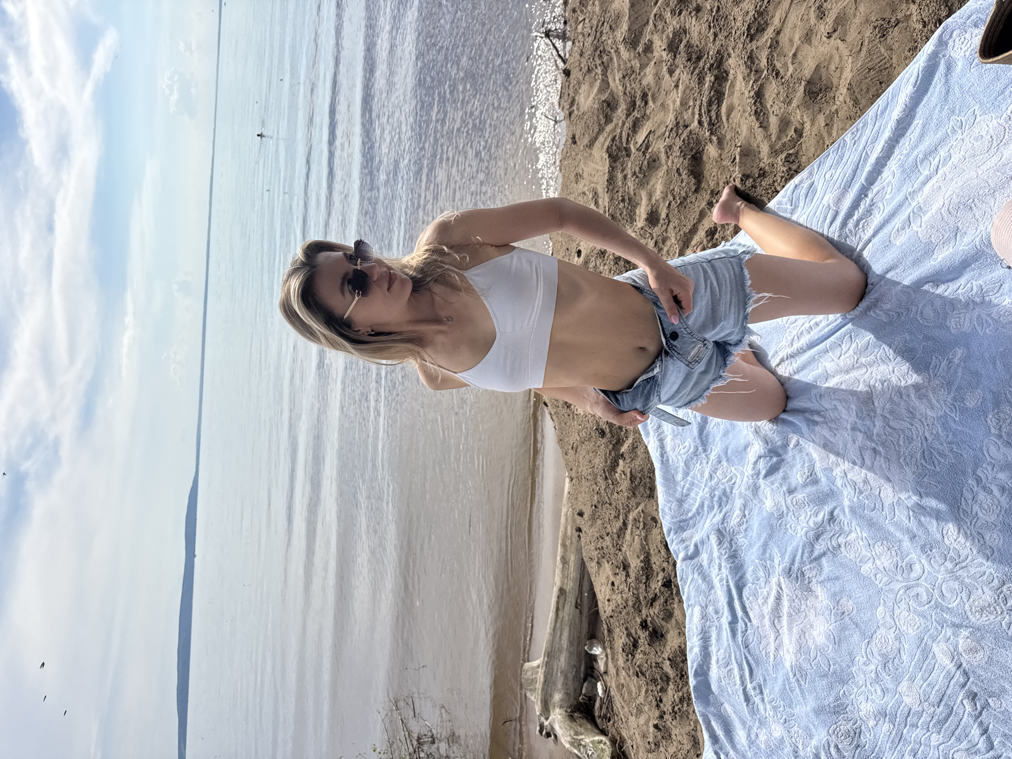 A woman is kneeling on a light blue towel on a sandy beach. She is wearing a white bikini top and denim shorts. She has blonde hair and is wearing sunglasses. Her hands are on her shorts. The beach is next to a body of water, and the sky is cloudy. The sun is shining, indicated by the shadows. The scene appears to be taking place during the daytime. There is a piece of driftwood on the sand next to the woman.