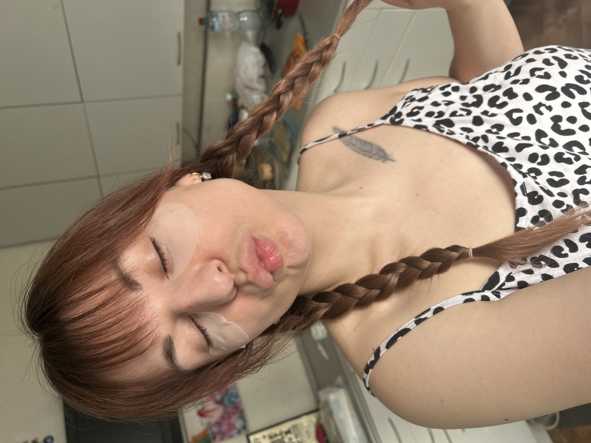 The image depicts a female individual indoors, in what appears to be a kitchen or utility area in the city of Foky, Russia. The subject has auburn hair styled in bangs across her forehead and two long braids extending over her shoulders. She wears translucent eye patches beneath both closed eyes and has her lips pursed in a pouting expression. Gold-colored hoop earrings with clear stones are visible in both ears. She is dressed in a white camisole featuring a black leopard print, and a black feather tattoo is visible on her left clavicle. Her right hand is positioned near the end of the braid on the right side of the frame. The background includes white cabinetry with horizontal lines, a white countertop, and various items such as a plastic water bottle, a red pot, and other indistinct objects.