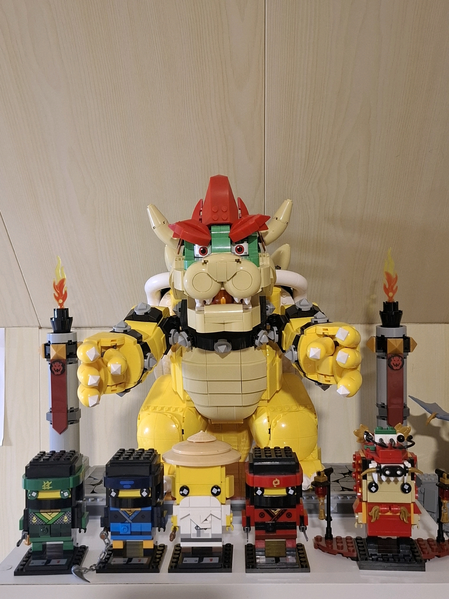 This is a medium shot, eye-level photograph of a collection of LEGO figures displayed on a shelf against a light wood-paneled wall. Dominating the scene is a large, yellow, Bowser-like LEGO character with a red headdress, green eyes, and spiked arms. In front of Bowser, arranged from left to right, are five smaller LEGO figures: a green ninja with a black mask, a blue ninja with a black mask and a chain weapon, a yellow figure with a straw hat and white clothing, a red ninja with a black mask and red clothing, and a red and gold figure with a headdress and a mustache. Flanking Bowser are two cylindrical LEGO structures with flame elements at the top. The overall impression is one of a well-arranged display of popular culture-themed LEGO creations.