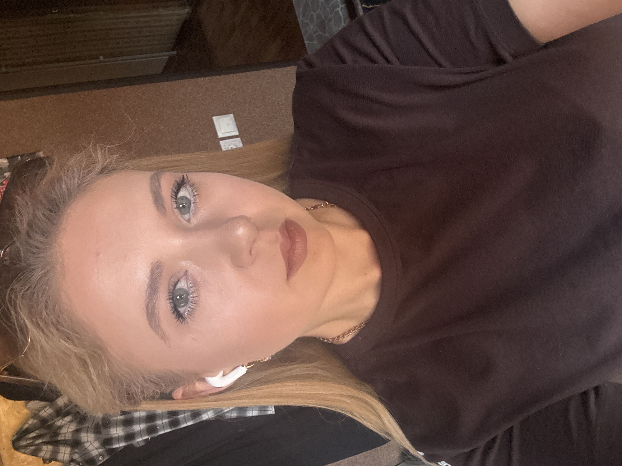 A medium shot shows a woman with blonde hair, fair skin, and blue eyes. She is wearing a dark brown t-shirt and white earbuds. She is also wearing a gold chain necklace. She is looking directly at the camera. The background is a textured brown wall with two light switches. A plaid shirt is visible on the left side of the image. The overall lighting is warm, with shadows suggesting an indoor setting.