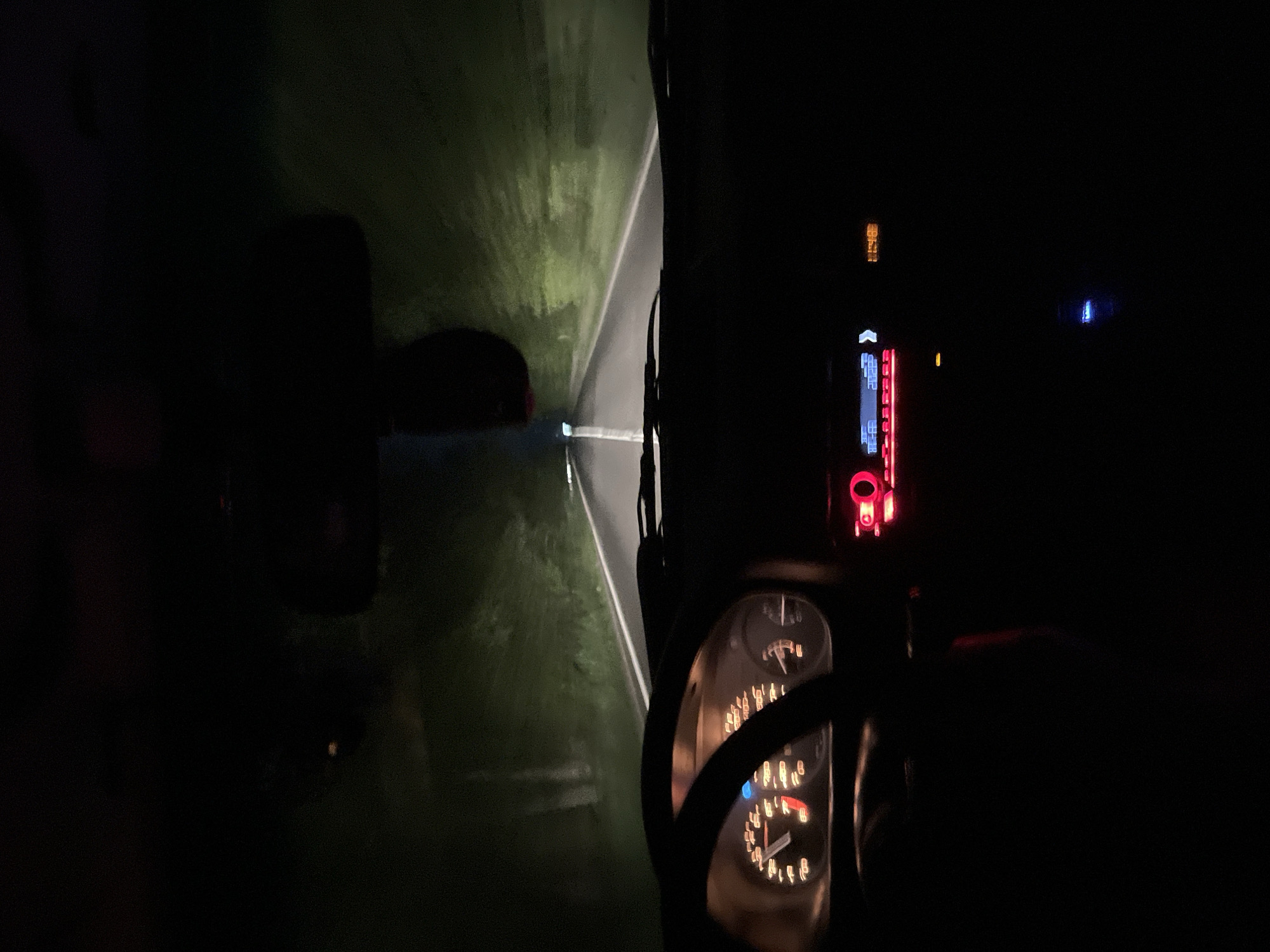 A nighttime interior view from a vehicle moving on a rural road. The road is illuminated by the vehicle's headlights, showing the paved surface and grassy edges. The dashboard is visible, including gauges and an electronic display with red and blue illumination. The location is unknown as no details indicate Dibich, Bulgaria. No people are visible.