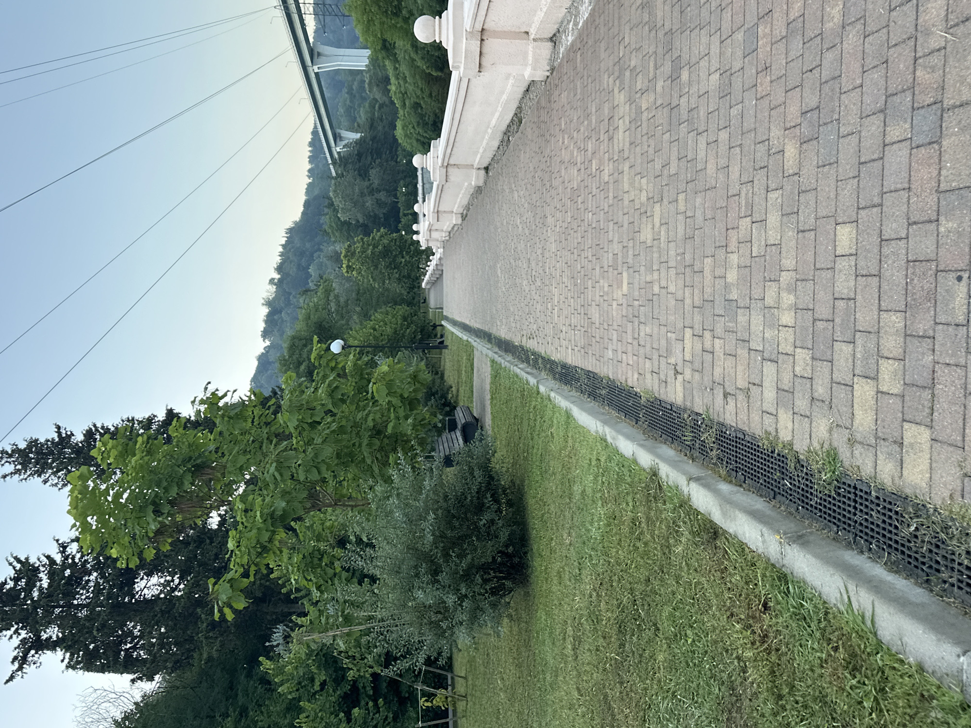 A paved pathway with a textured surface made of light brown and gray bricks leads into the distance. The pathway is bordered on the right by a cream-colored decorative wall with rounded finials on top and substantial square posts. On the left, the pathway is separated from a grassy area by a concrete curb and a black metal grate drain. The grassy area contains a dark wooden park bench and a lush green shrub. Trees with dense green foliage are visible behind the grassy area. In the background, a modern bridge with white concrete pillars spans across a green, tree-covered valley. Overhead, several thick electrical wires are suspended in the pale blue sky. The scene appears to be a park or promenade in Sochi, Russia, likely near a body of water or elevated area due to the presence of the bridge. The lighting suggests it is daytime, possibly early morning or late afternoon.